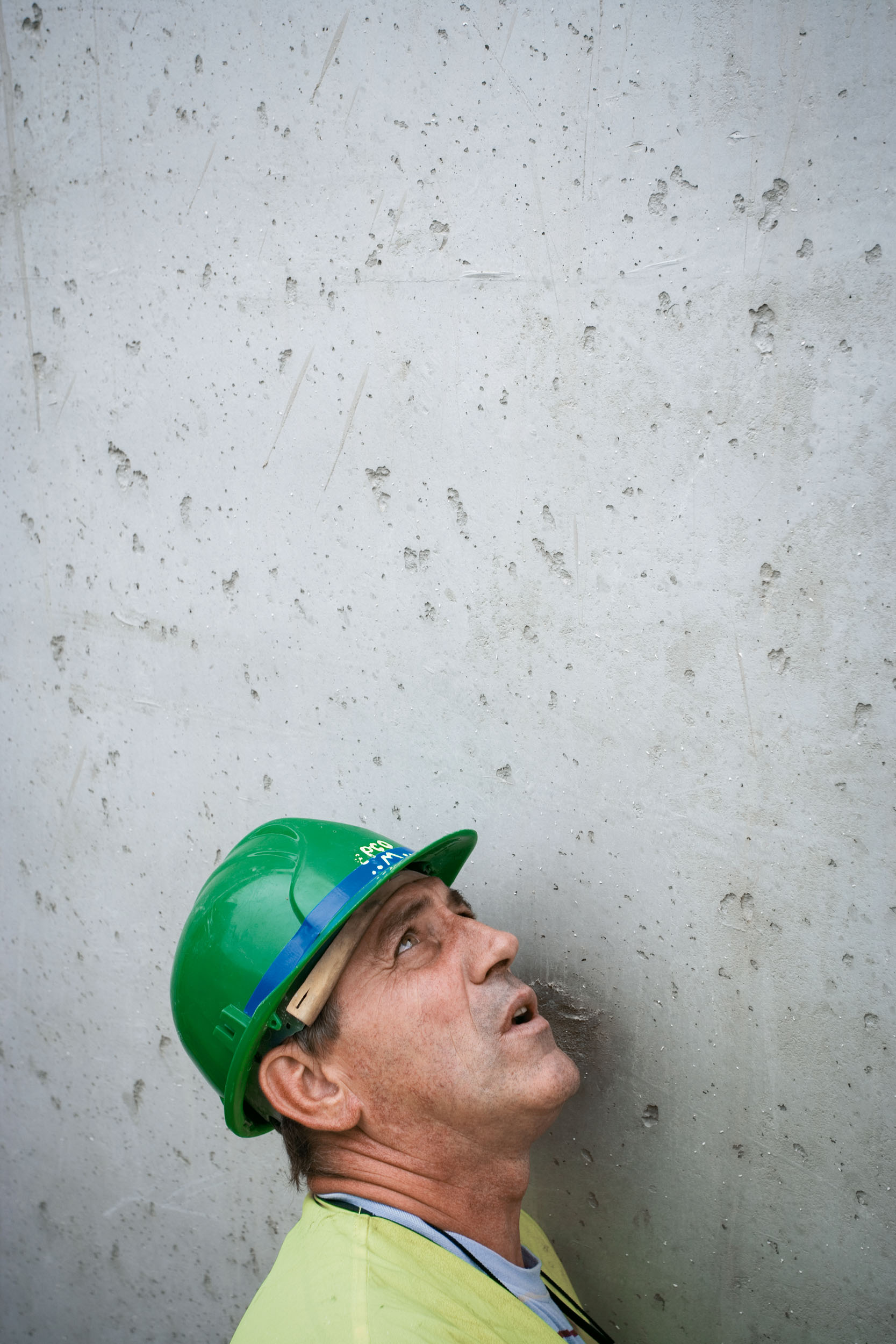 Photo de portrait d'un ouvrier casqué sur un fond de mur en béton fraîchement coulé, il est de profil, collé au mur, et regarde attentivement vers le haut