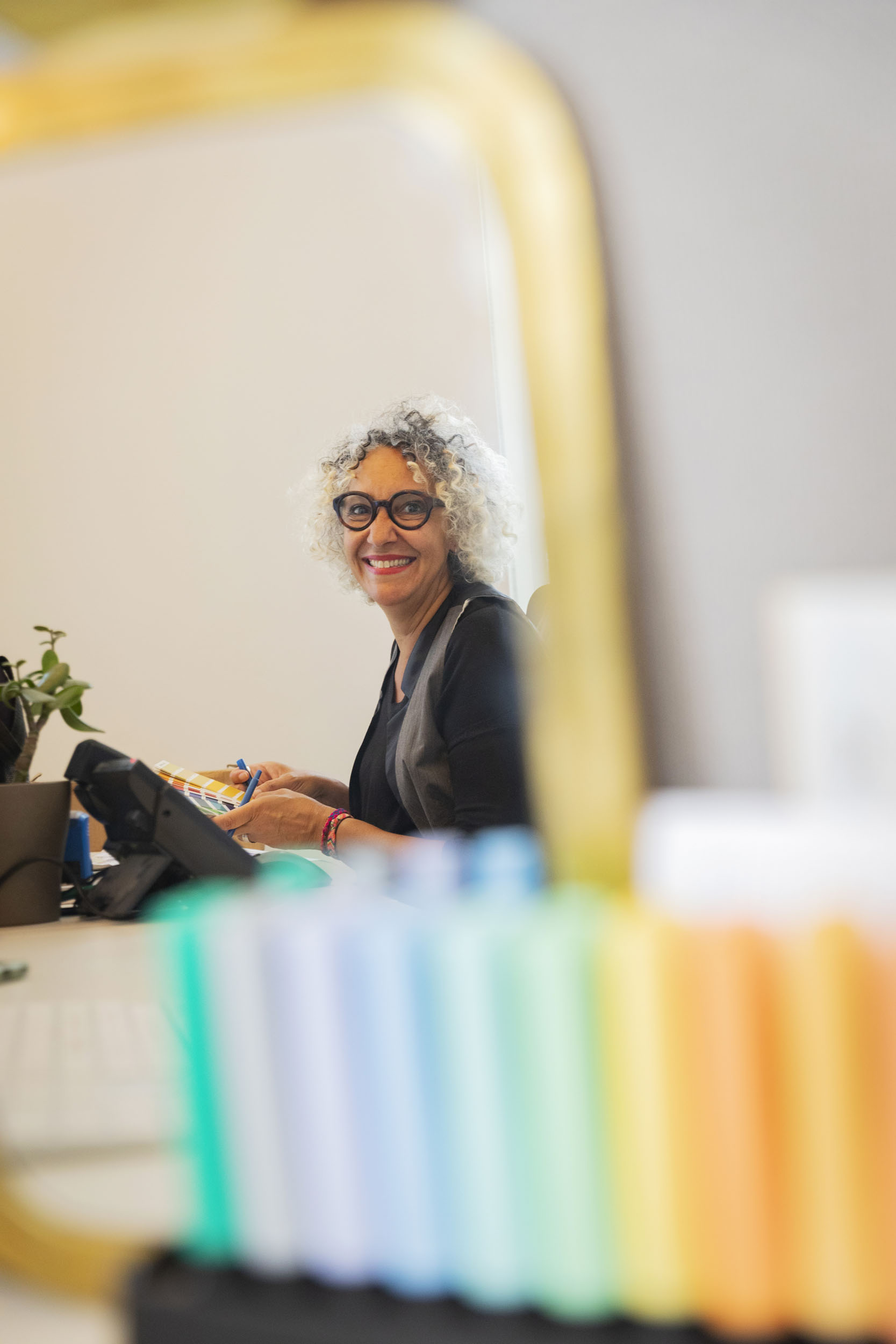 Portrait dans un miroir d'une femme souriante au travail à son bureau tenant un nuancier de couleurs Pantone à la main, elle regarde dans l'objectif