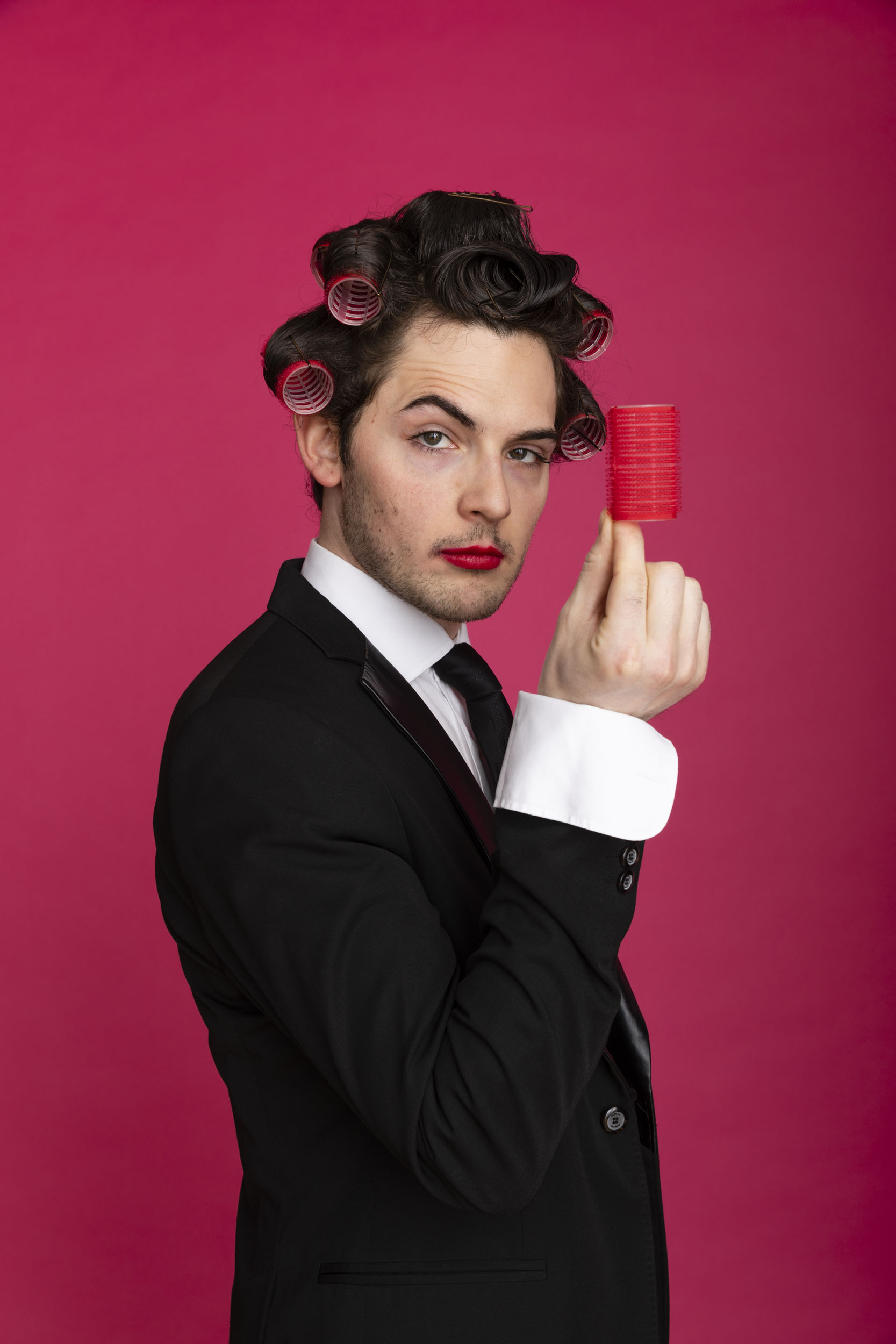 Portrait d'un homme en costume et cravate noir sur fond rose qui regarde dans l'objectif. Il porte des bigoudis dans les cheveux, du rouge à lèvres, et tient un bigoudi dans sa main droite