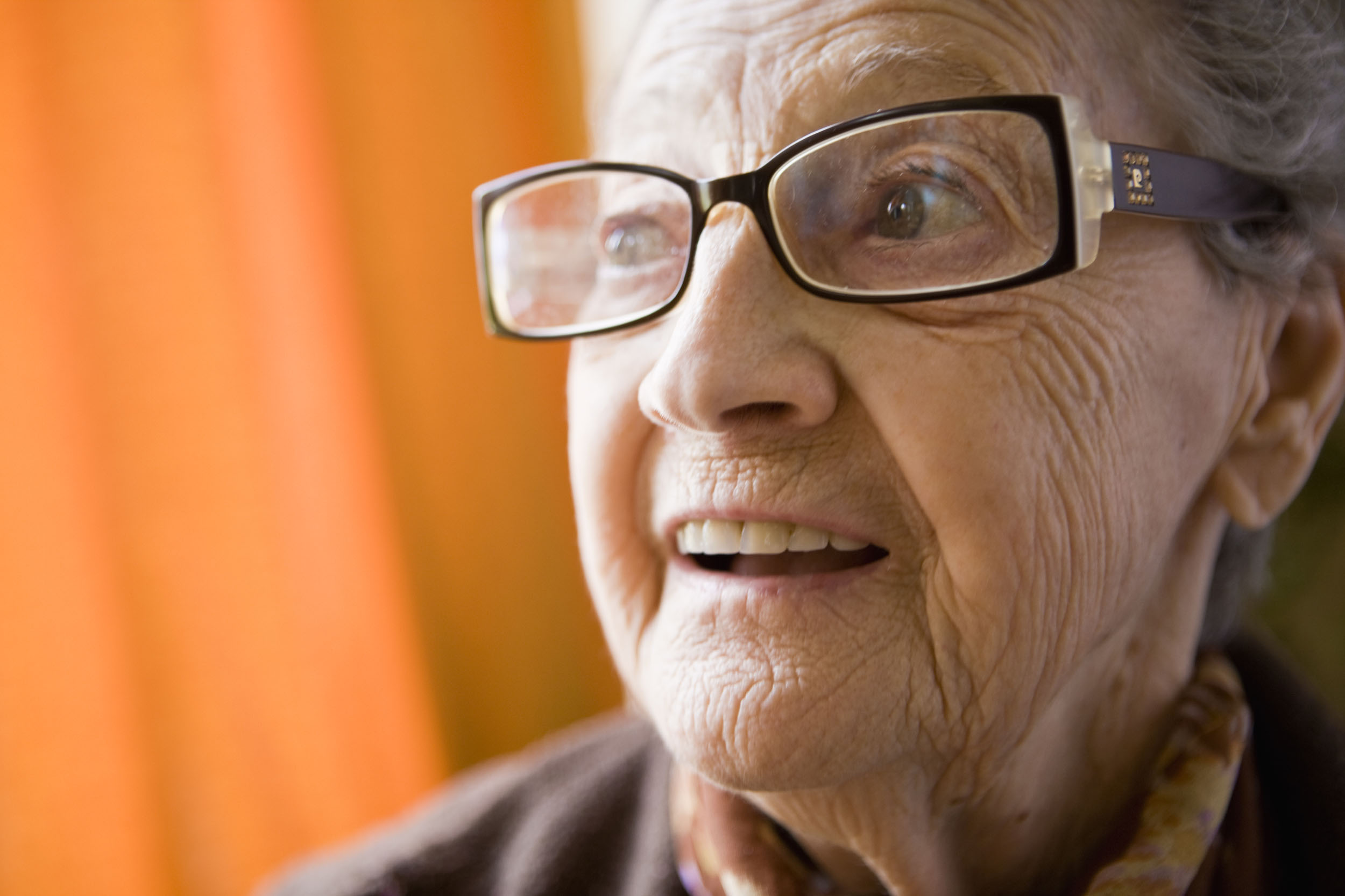 Photo de portrait en gros plan d'une femme âgée ridée avec des lunettes sur fond orange. Elle a la bouche ouverte et le regard dans le vague.