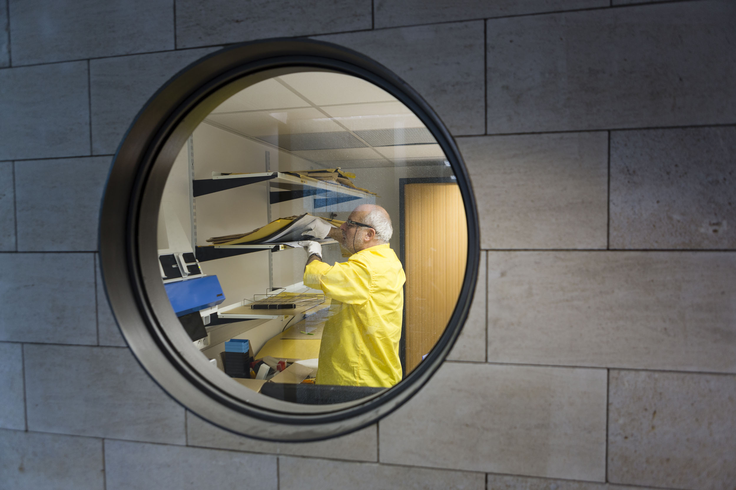 Vue à travers une vitre ronde intégrée dans le mur d'un homme au travail vêtu d'une chemise jaune cherchant des clichés sur une étagère