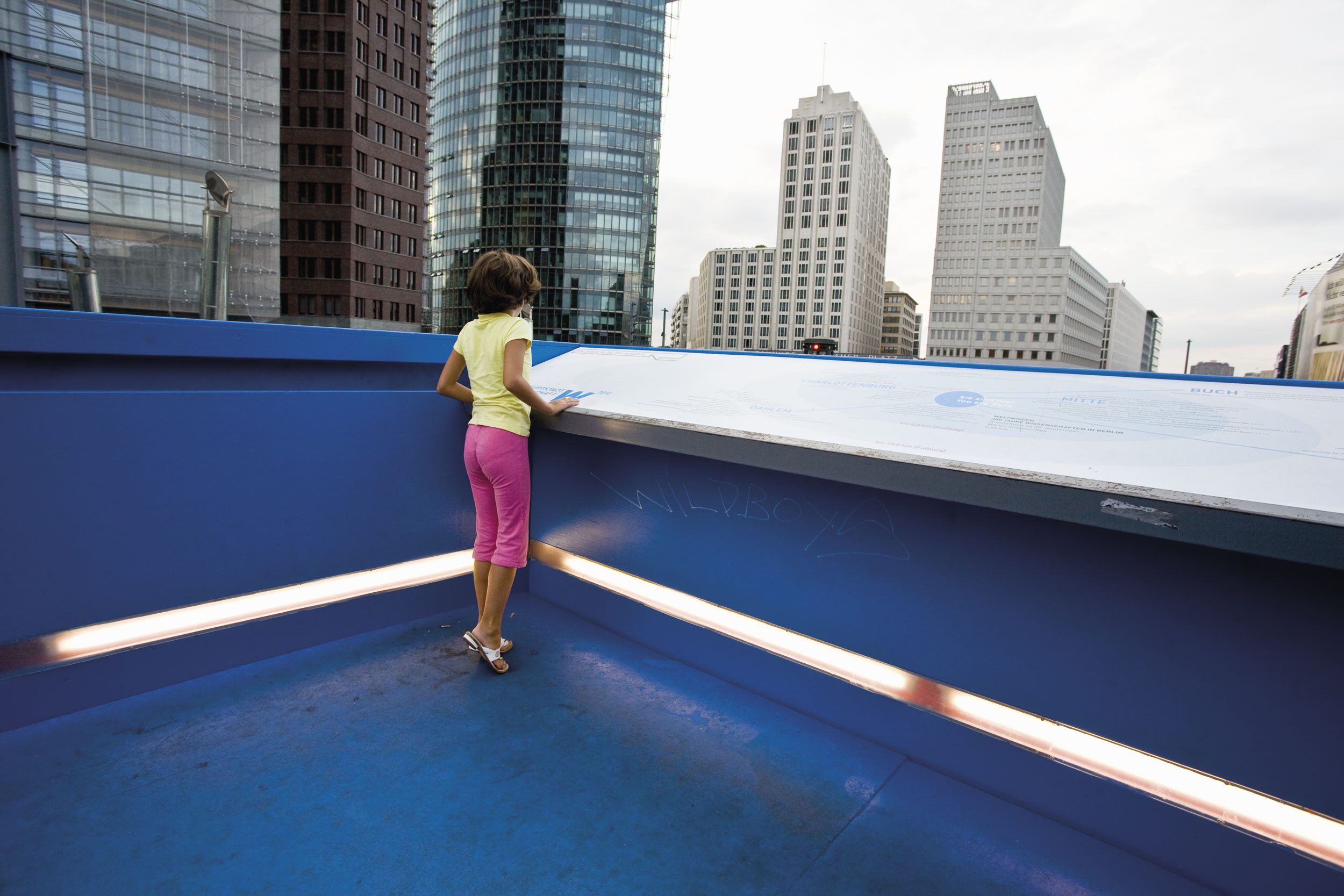 Jeune fille de dos debout portant un t-shirt jaune et un pantalon rose juchée sur un promontoire bleu, elle regarde la vue sur les grands immeubles devant elle à Berlin