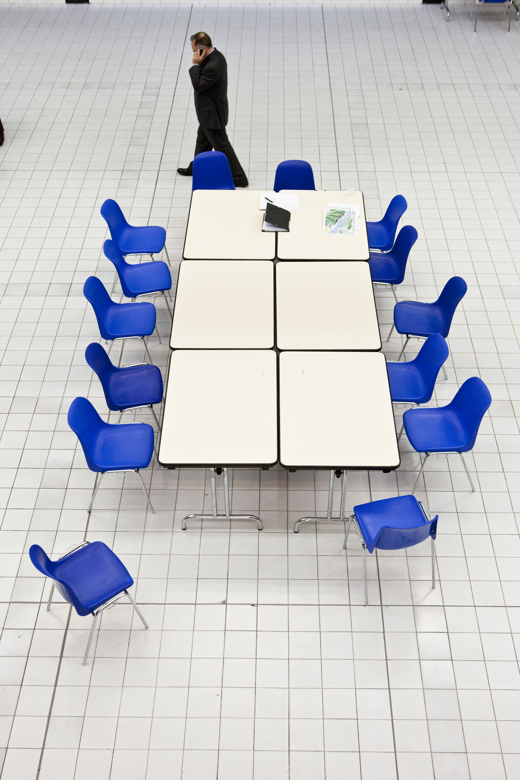 Un homme seul téléphone en marchant dans une salle de réunion vide au carrelage blanc, 6 tables blanches sont au centre entourées de 14 chaises en plastique bleu