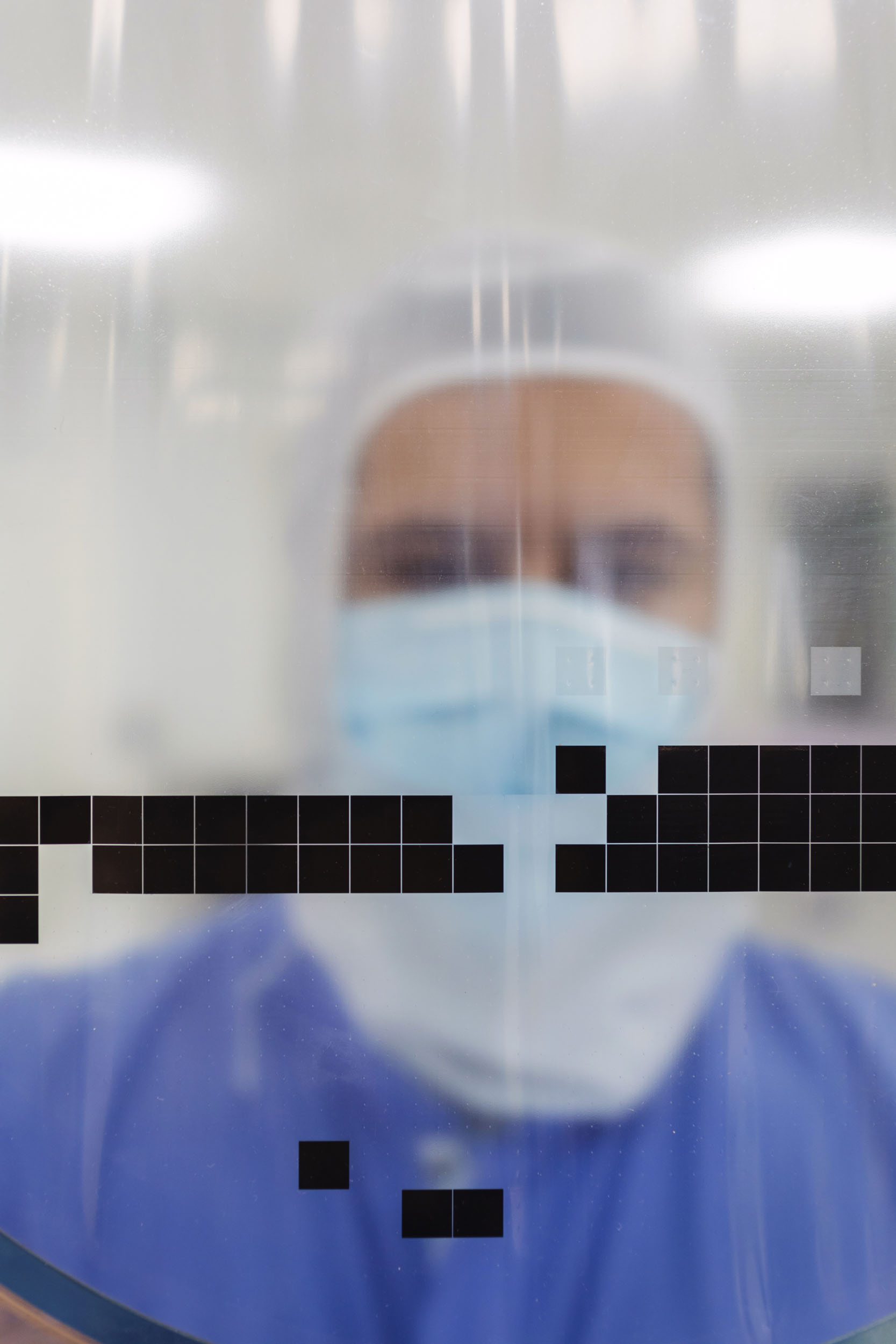 Portrait d'un homme dans une salle blanche vu à travers une matière plastique translucide sur laquelle on distingue des éléments carrés noirs nets au premier plan, son visage à l'arrière plan est flou