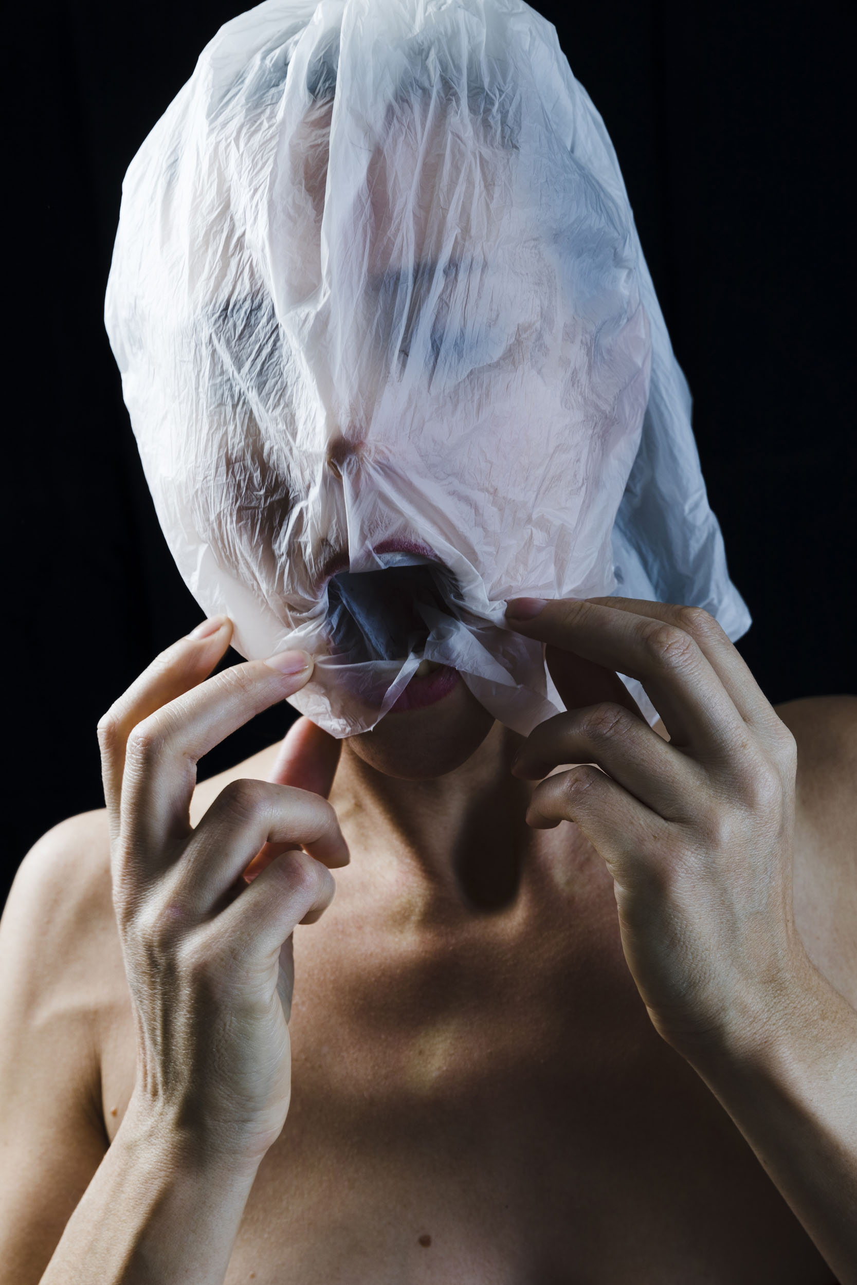 Femme torse nu sur fond noir qui s'est enfilée un sac en plastique translucide sur la tête, elle inspire les yeux fermés et maintient le sac avec ses mains