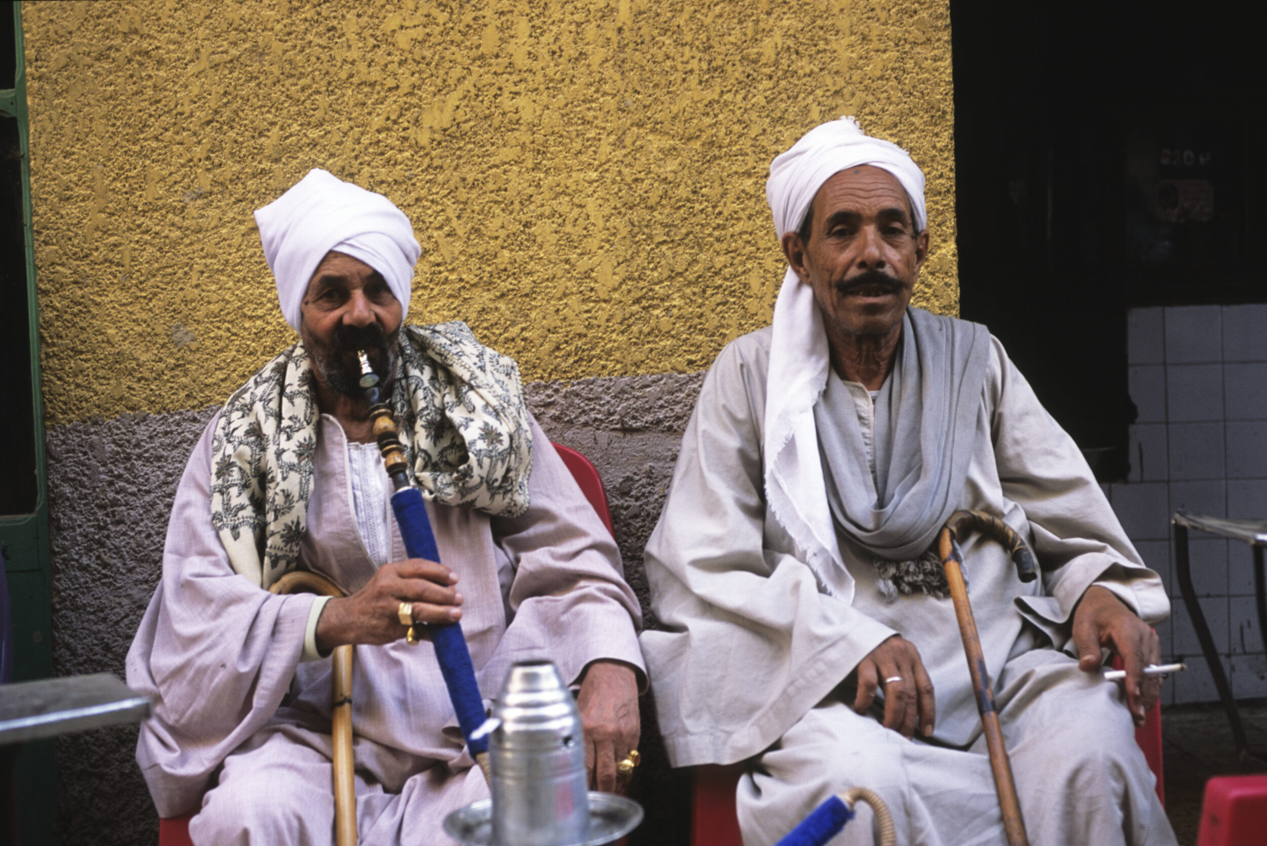 2 hommes en djellaba et cheikh sont assis dans une rue du quartier de Mokattam au Caire en Égypte, celui de gauche fume le narguilé, l'homme à droite fume une cigarette