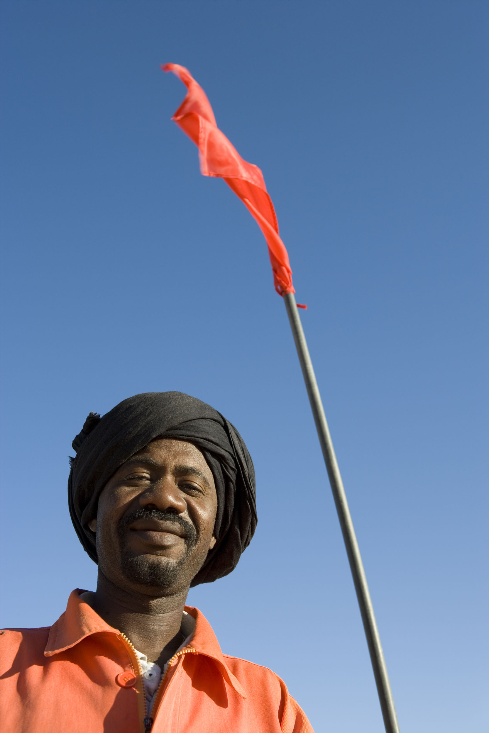 Portrait en contre plongée d'un homme souriant au travail portant un cheich noir sur la tête. Il est vêtu d'une veste orange. Un drapeau rouge flotte au dessus de sa tête, le ciel bleu