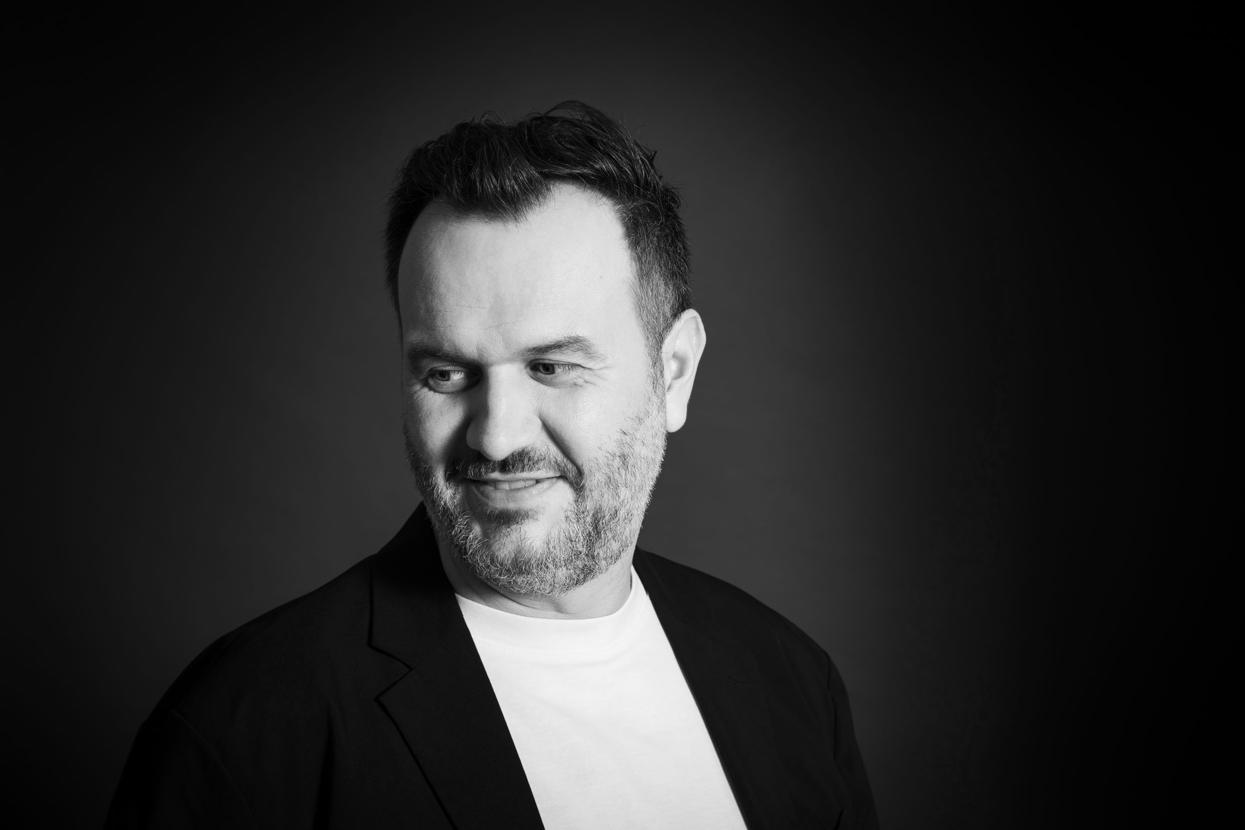 Portrait de profil réseau sociaux en noir et blanc d'un homme en t-shirt blanc et veste sombre cadrée en plan buste, il porte la barbe et la moustache et regarde vers le sol