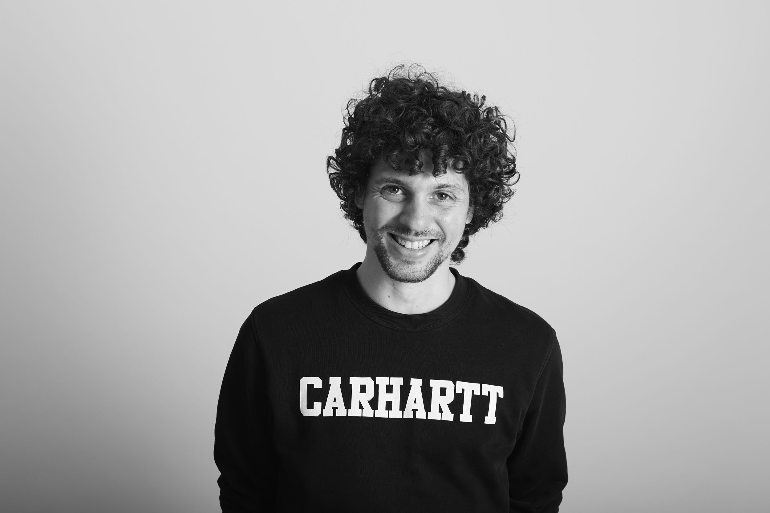 Portrait de profil réseau social en studio sur fond gris clair d'un homme souriant et décontracté aux cheveux bouclés portant en sweat-shirt
