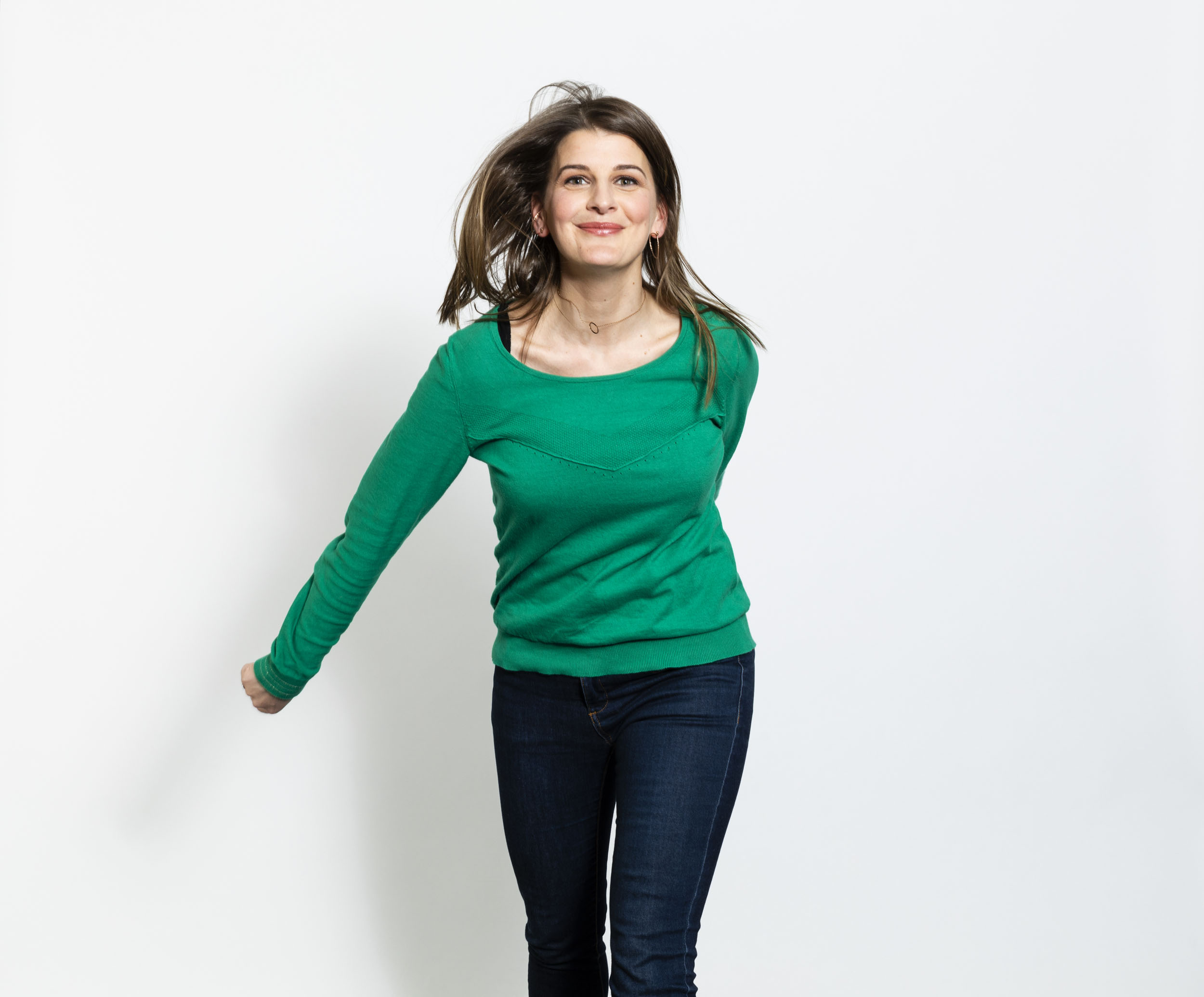 Portrait de profil réseau sociaux en couleurs d'une femme en jeans et pull vert qui saute en regardant dans l'objectif, portrait réalisé en studio sur fond blanc