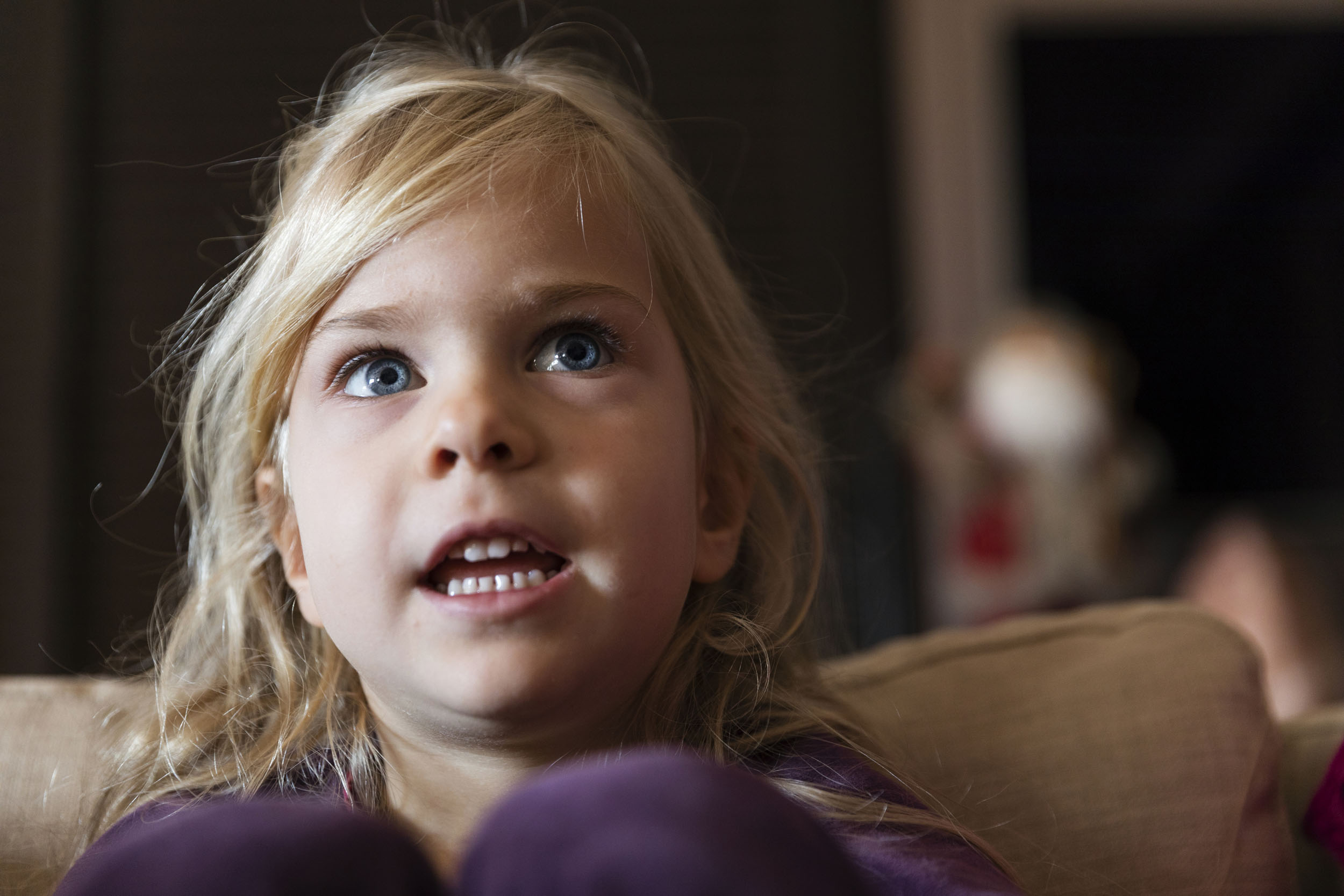 Portrait d'une jeune enfant blonde aux yeux bleus captivés. Elle est assise dans un canapé et a la bouche ouverte