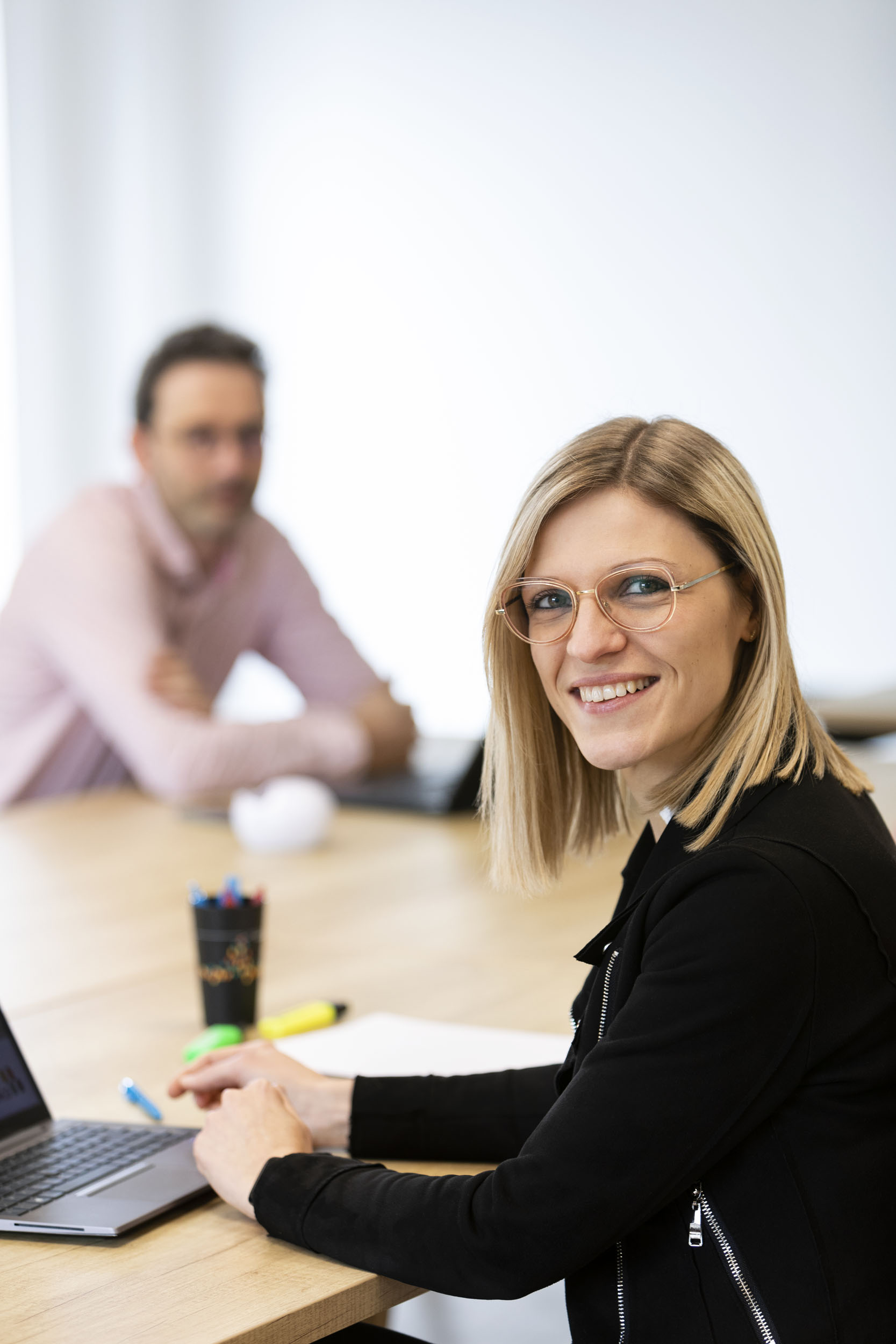 Portrait d'une femme blonde et souriante en réunion, elle porte un vêtement noir et regarde l'objectif. Un homme est flou en arrière plan à gauche