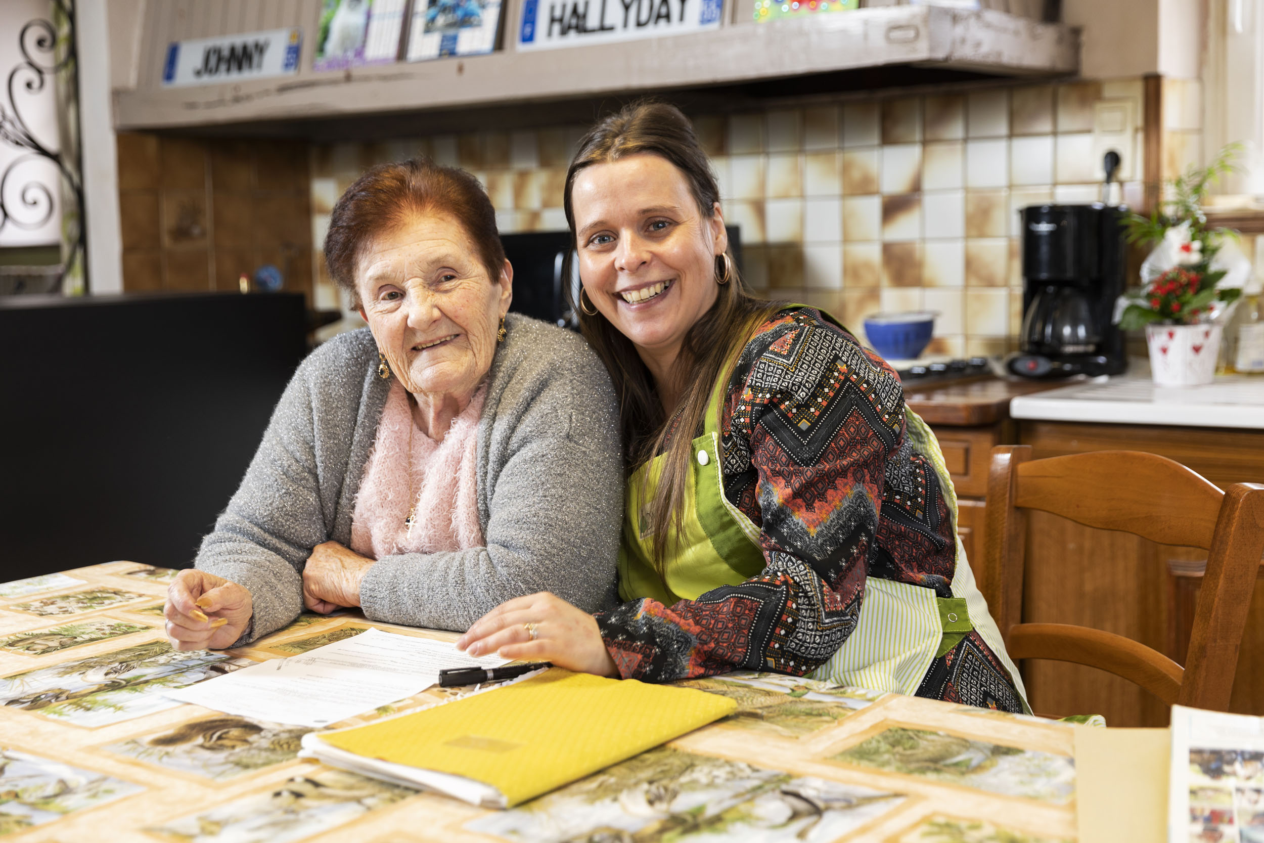 Portrait d'une jeune aide ménagère au domicile d'une dame âgée dans la Meuse. Elles sont souriantes et assises côte à côte à la table de la cuisine recouverte d'une toile cirée
