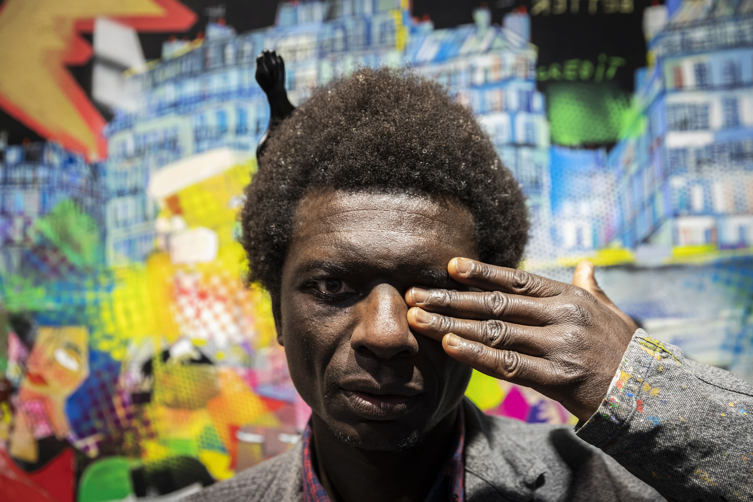Portrait de l'artiste peintre Olivier Anicet devant une de ses œuvres dans une galerie Carré d'Artistes à Paris, il porte une blouse grise et cache son œil gauche avec sa main gauche