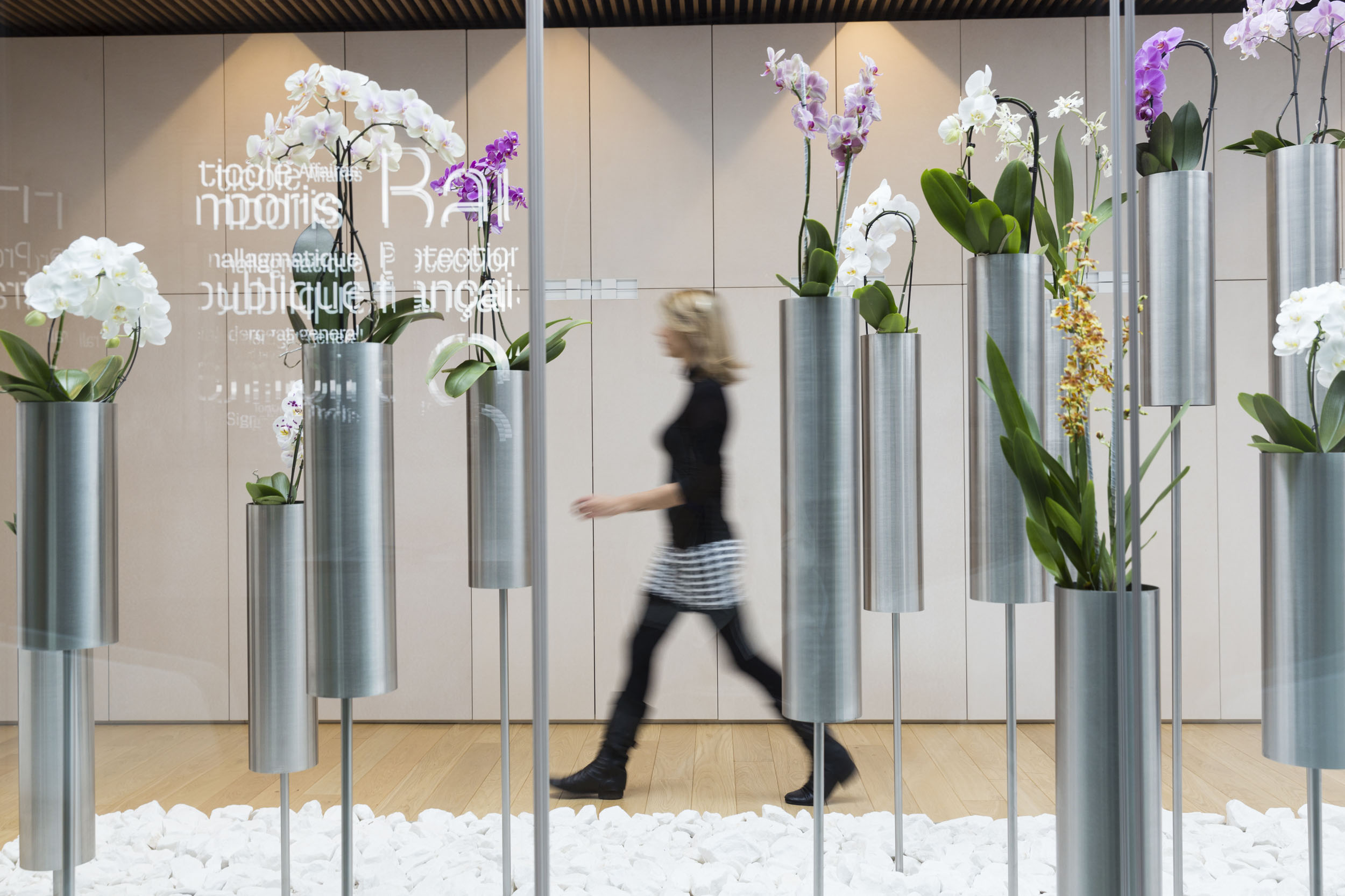 Portait artistique de plain-pied d'une femme blonde en jupe à damier noir et blanc et pull noir marchant derrière des orchidées dans un intérieur design