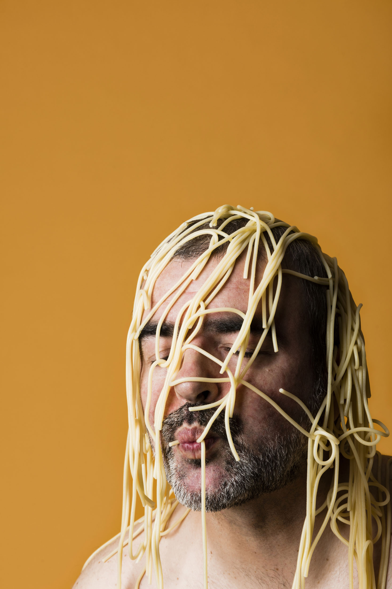 Portrait artistique en gros plan sur fond jaune orange d'un homme torse nu les yeux fermés avec des spaghettis cuits sur la tête, il aspire un spaghetti avec sa bouche