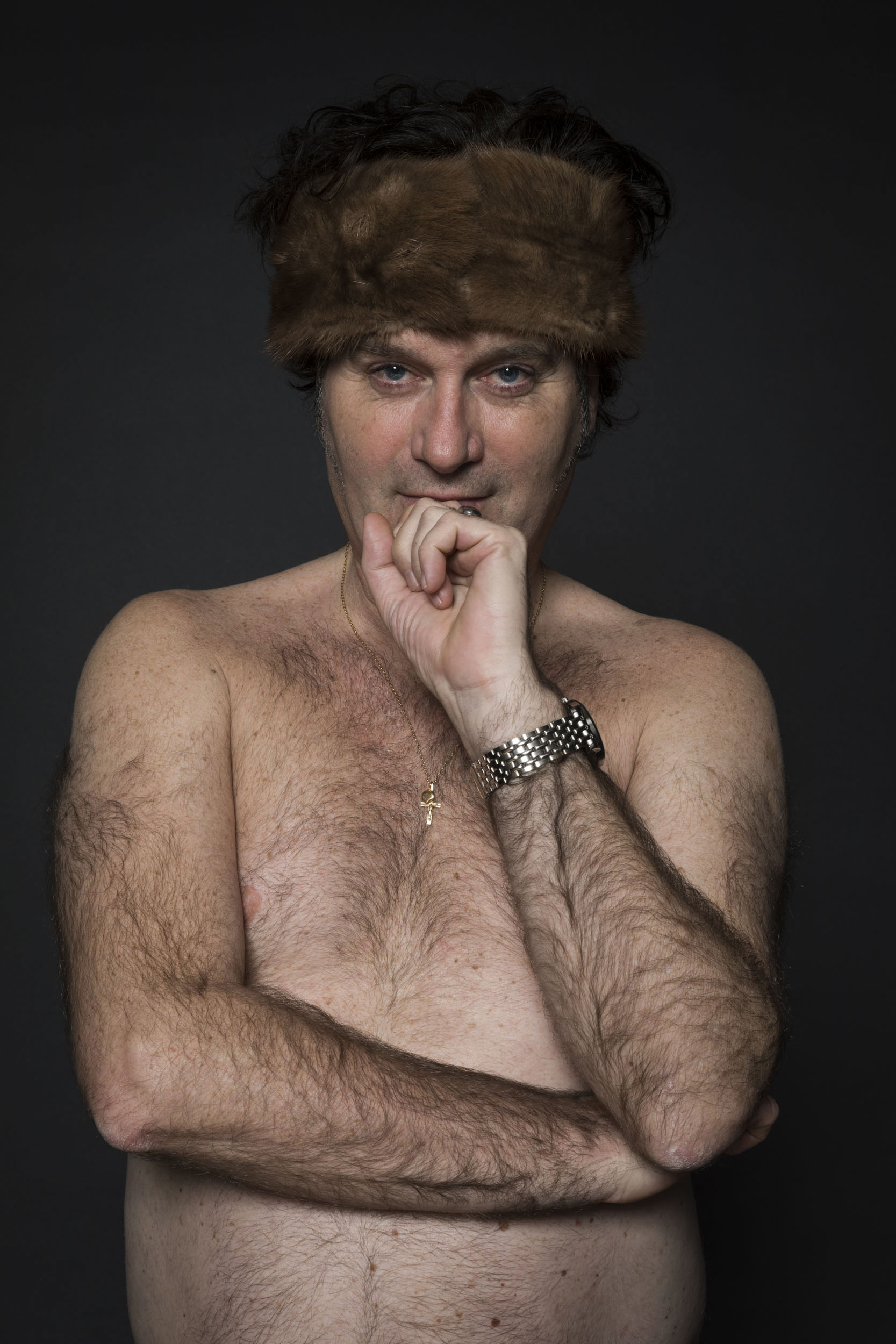 Portrait artistique d'une homme poilu et torse nu au regard narquois. Il porte une chaine avec une croix autour du cou et un bandeau en fourrure autour de la tête. Cadrage plan taille en studio sur fond noir