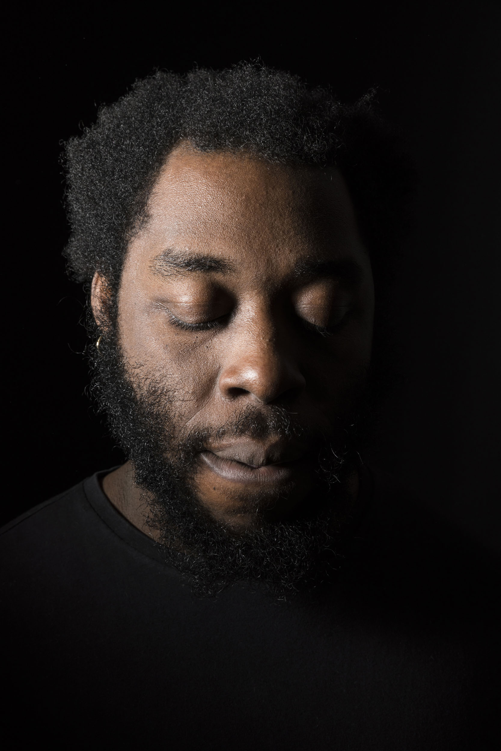 Portrait artistique d'un homme noir les yeux fermés sur fond noir qui porte la barbe et la moustache, le portrait est cadré en gros plan
