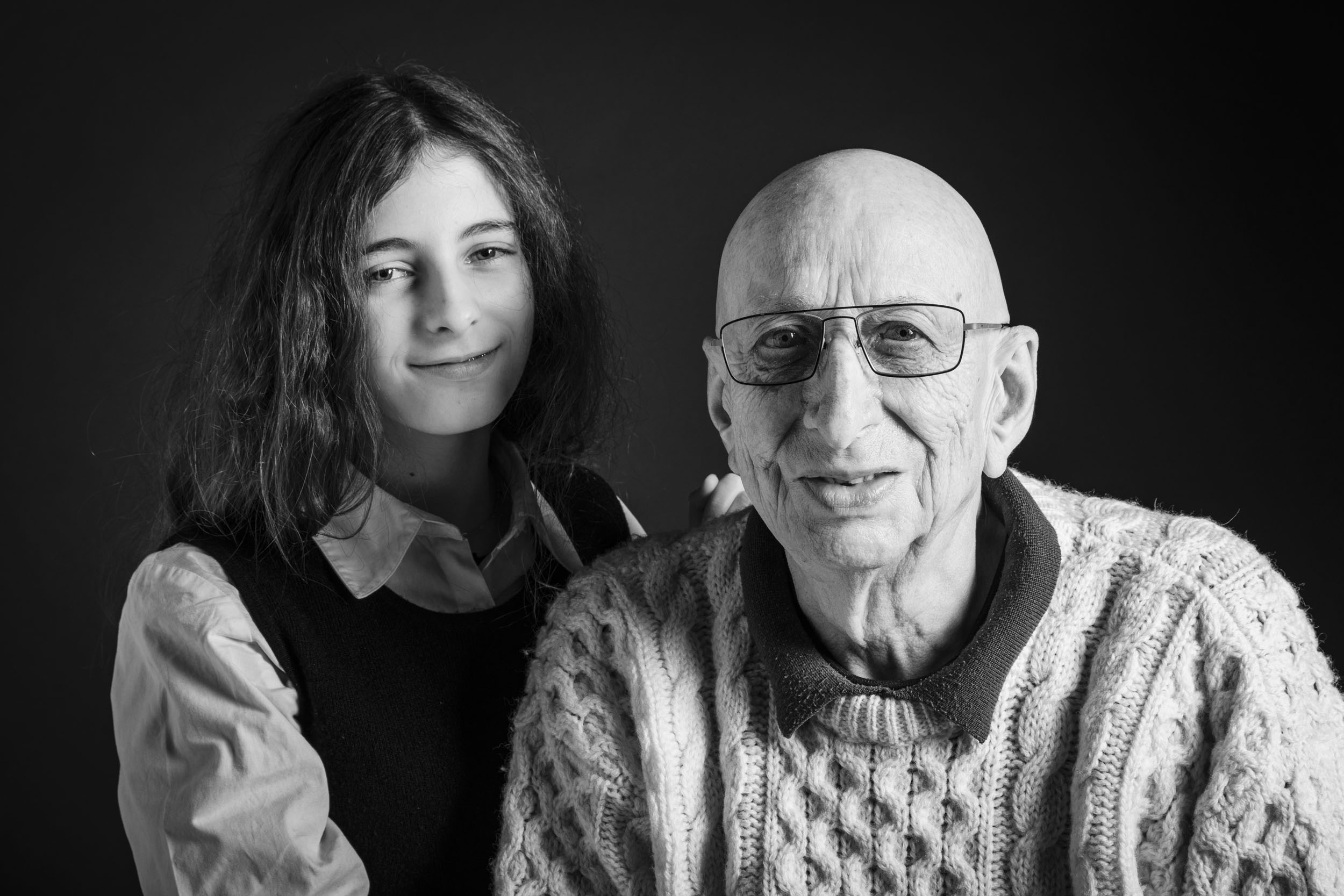 Portrait de famille en noir et blanc, un grand père portant un pull blanc avec sa petite fille souriante. Portrait en studio sur fond noir, cadrage en plan rapproché