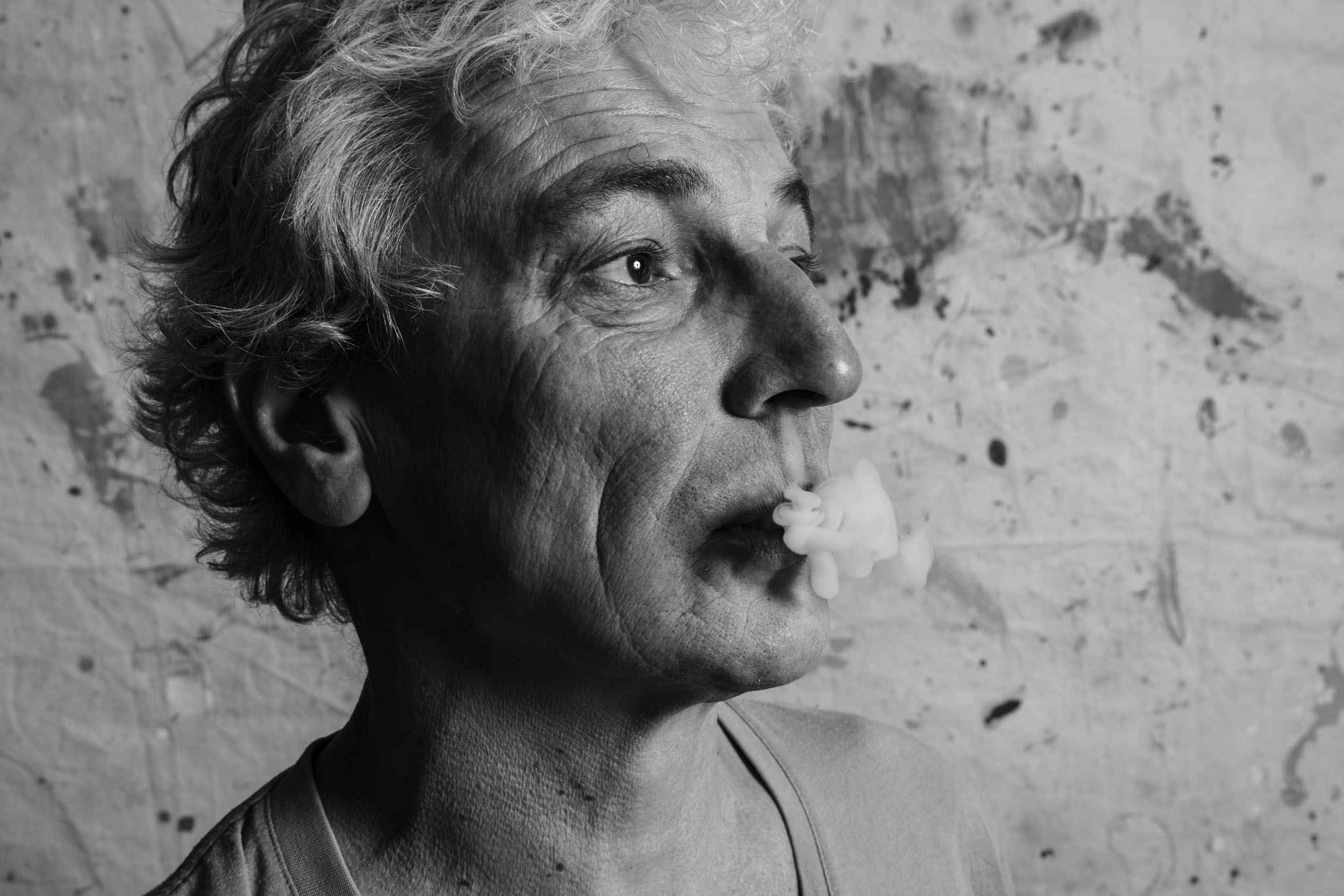 Portrait artistique en noir et blanc d'un homme en studio sur fond de bâche peinte. Il souffle de la fumée par le nez et regarde au loin