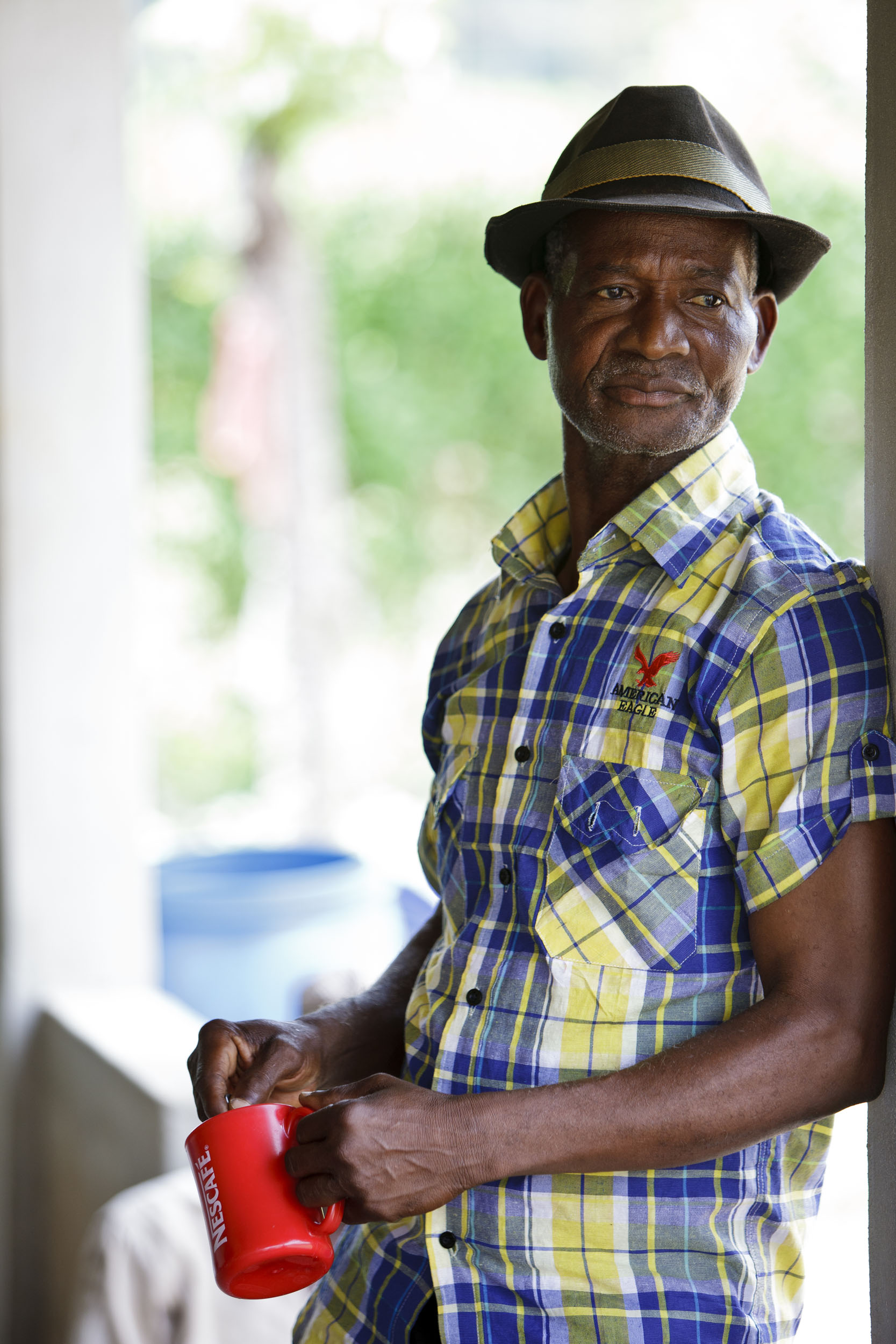 Portrait artistique d'un planteur de café adossé à un mur en Côte d'Ivoire, le regard dans le vague. Il porte un chapeau, une chemise à carreaux et tient un mug Nescafé® dans les mains