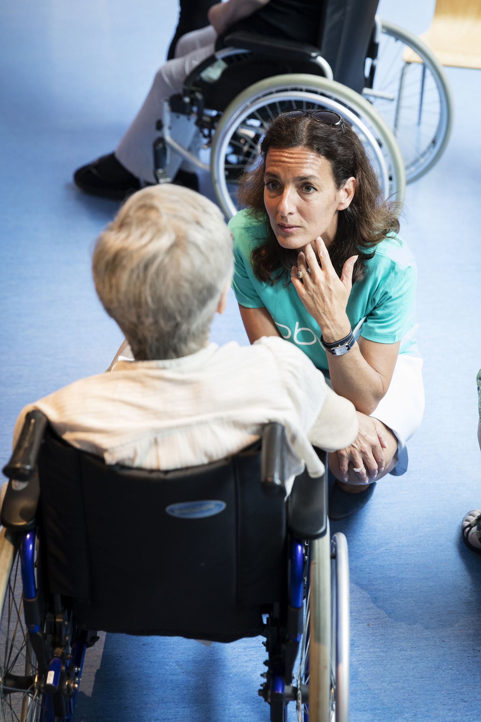 Portrait en plongée d'une femme bénévole qui échange avec une résidente assise de dos dans un fauteuil roulant après un récital de la chorale de l'entreprise pour l'Ehpah