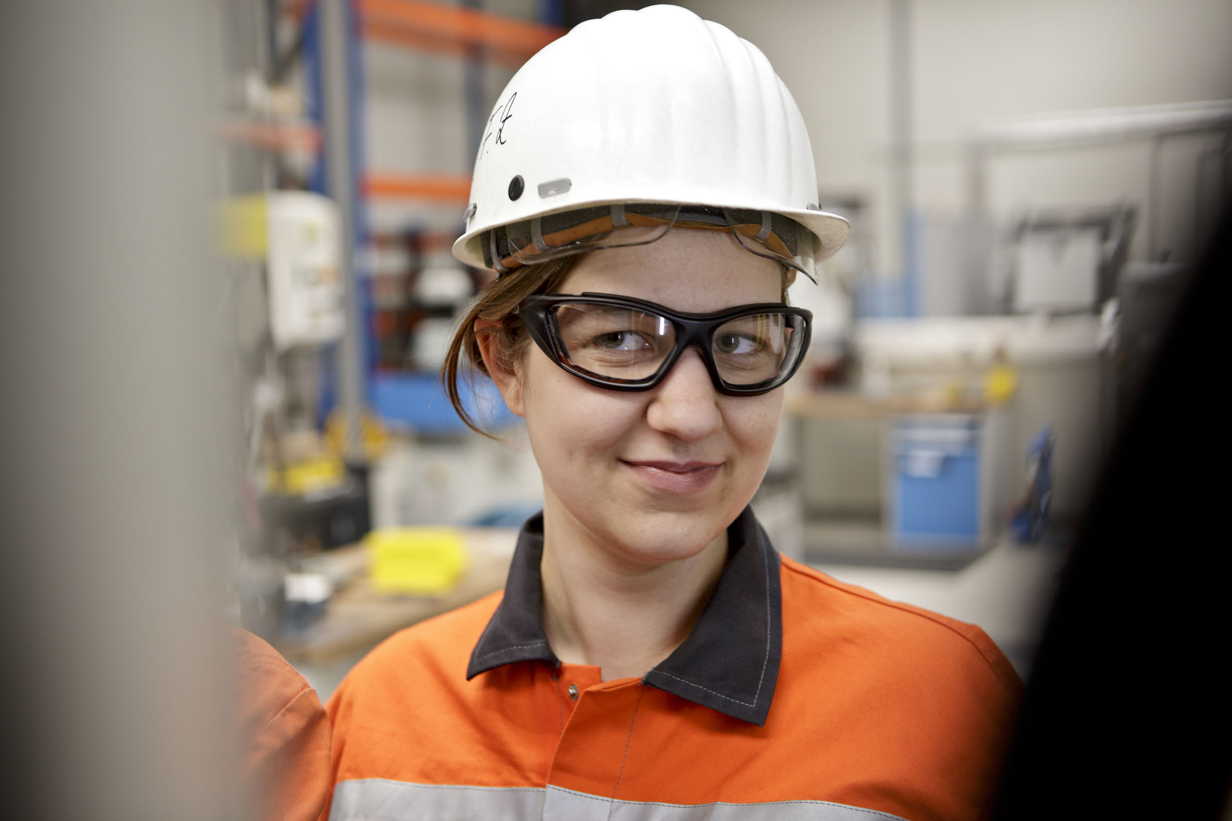 Portrait d'une jeune femme souriante portant un casque blanc, des lunettes de protection et une veste de travail orange à col marron dans un centre de recherche industriel