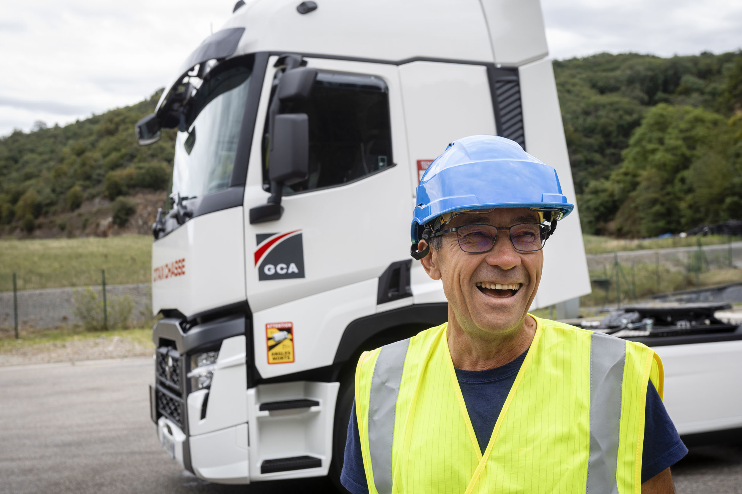 Portrait d'un chauffeur souriant debout devant son tracteur routier. Il porte un casque bleu et un gilet jaune fluorescent, le portrait est cadré en plan buste
