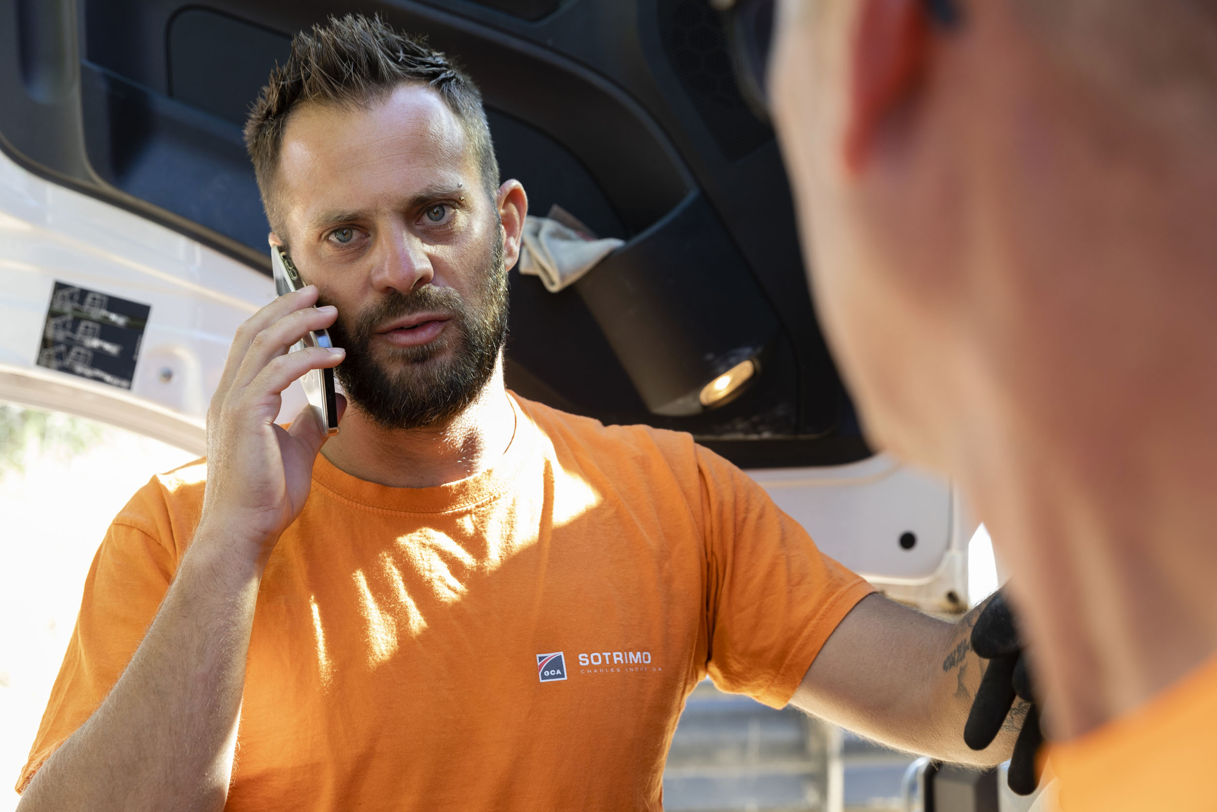 Portrait d'un chauffeur livreur portant la barbe et la moustache au téléphone devant son camion en arrière plan. Il porte un t-shirt orange