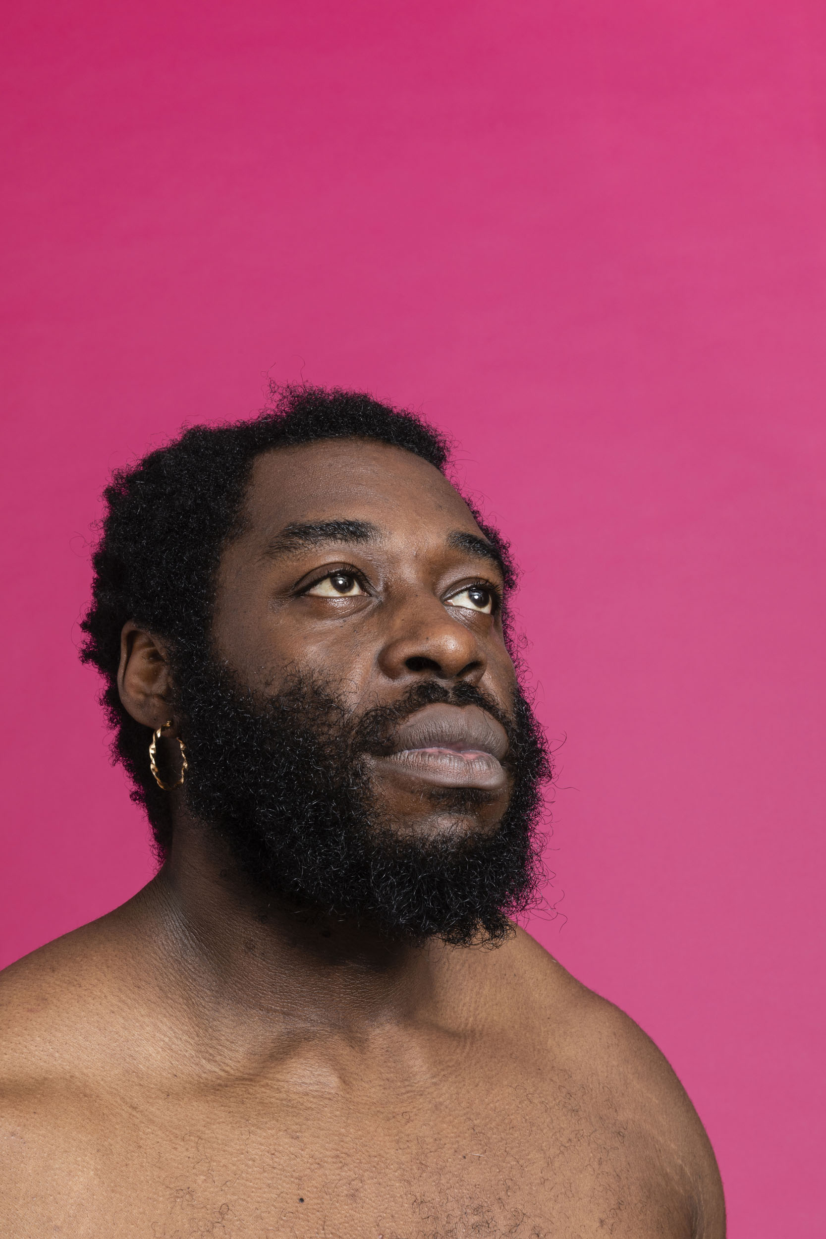 Portrait d'un homme noir barbu torse nu sur fond rose. Il regarde en hauteur sur sa gauche et porte un anneau à l'oreille droite