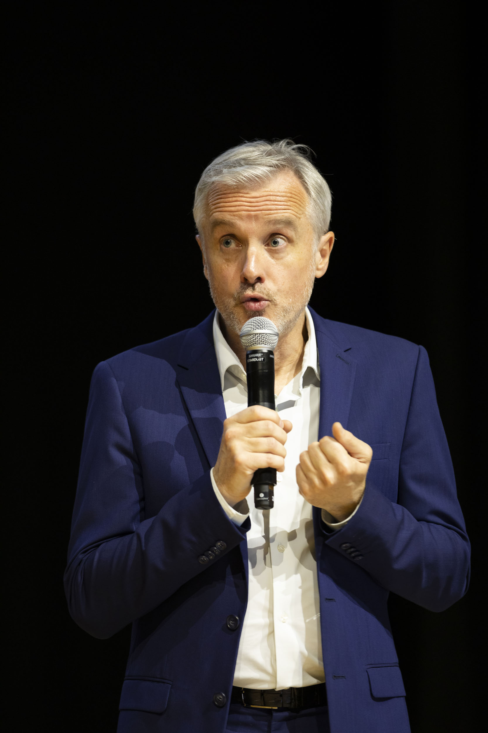 Photo de portrait d'un speaker sur scène en chemise blanche et veste bleu qui regarde le public en parlant dans le micro qu'il tient à la main lors d'une convention d'entreprise