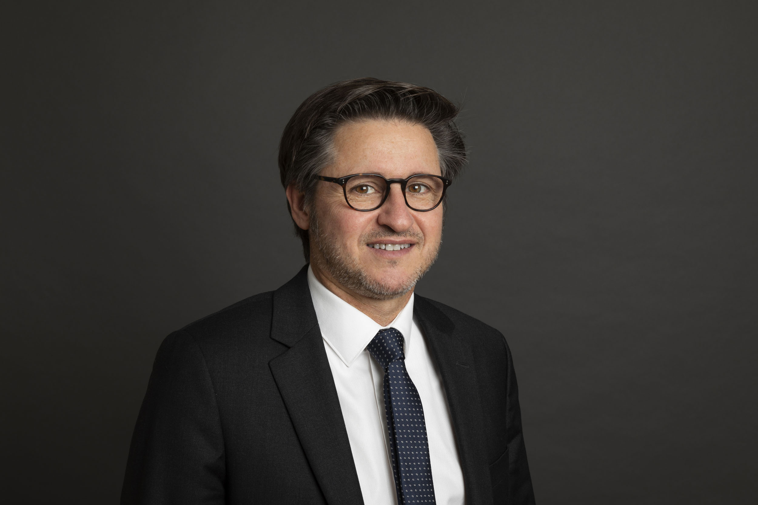 Photo de portrait d'un dirigeant souriant en studio avec des lunettes. Il porte une chemise blanche, une veste et une cravate, le fond est gris foncé