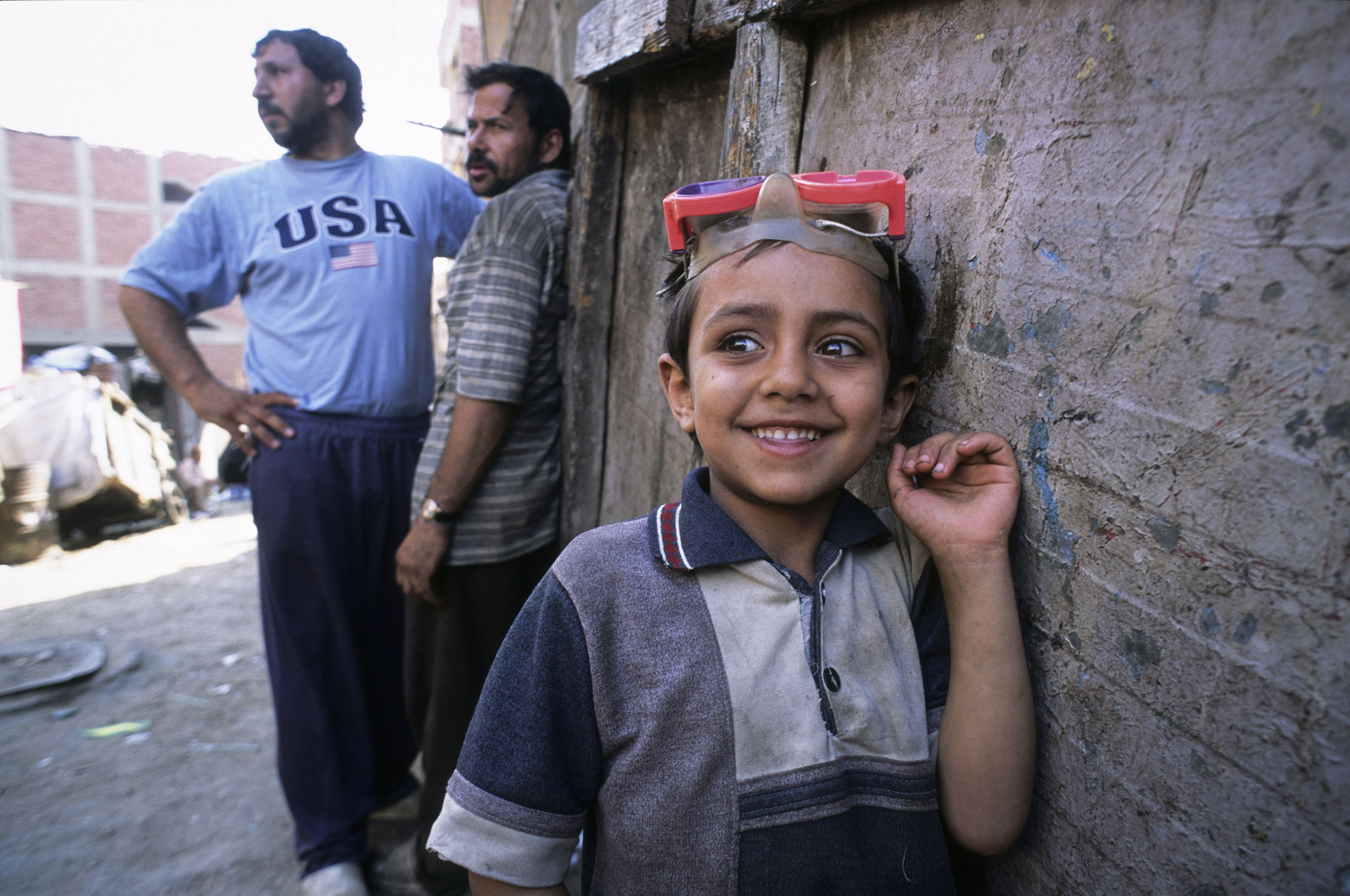 Portrait d'un enfant chiffonnier du Caire en Égypte, il est souriant et porte un masque de plongé sur la tête, 2 hommes se tiennent à l'arrière plan à gauche