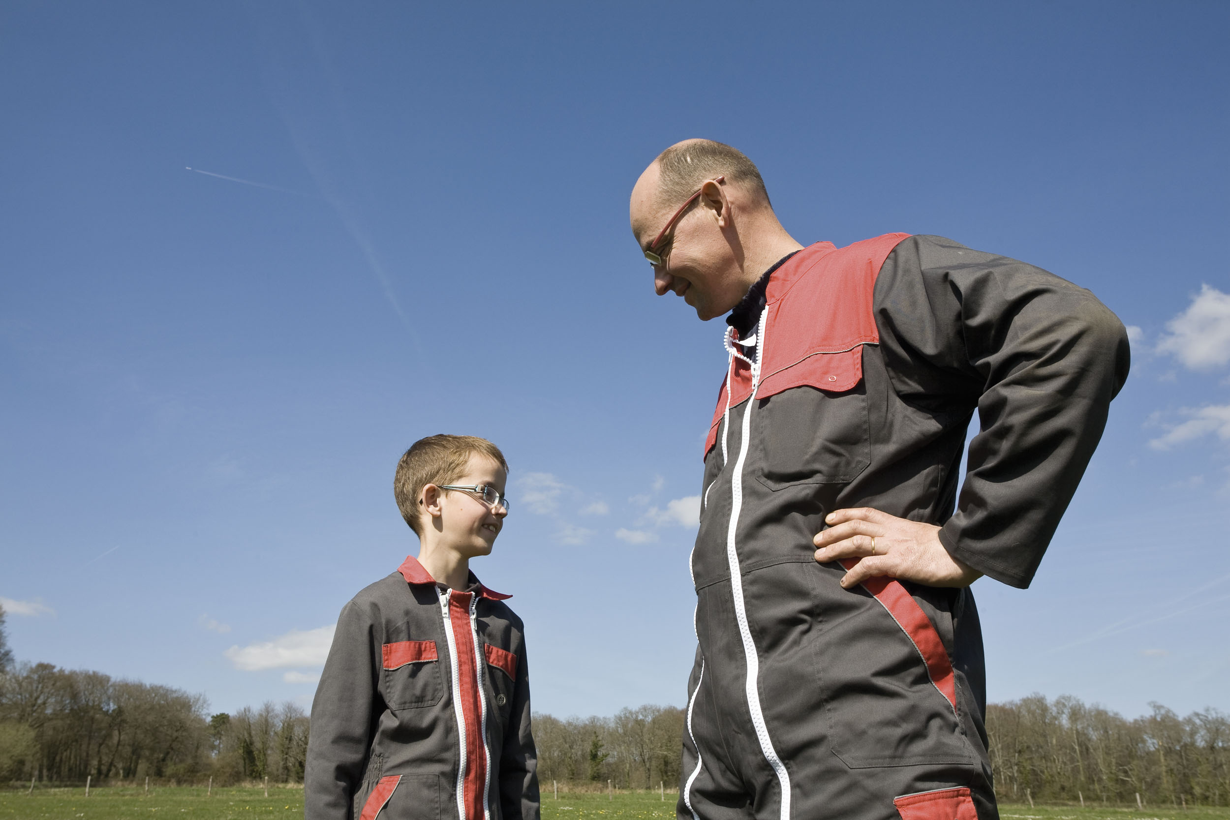 Portrait d'un père éleveur et son jeune fils debout côte à côte en combinaison de travail agricole dans une prairie sur fond de ciel bleu