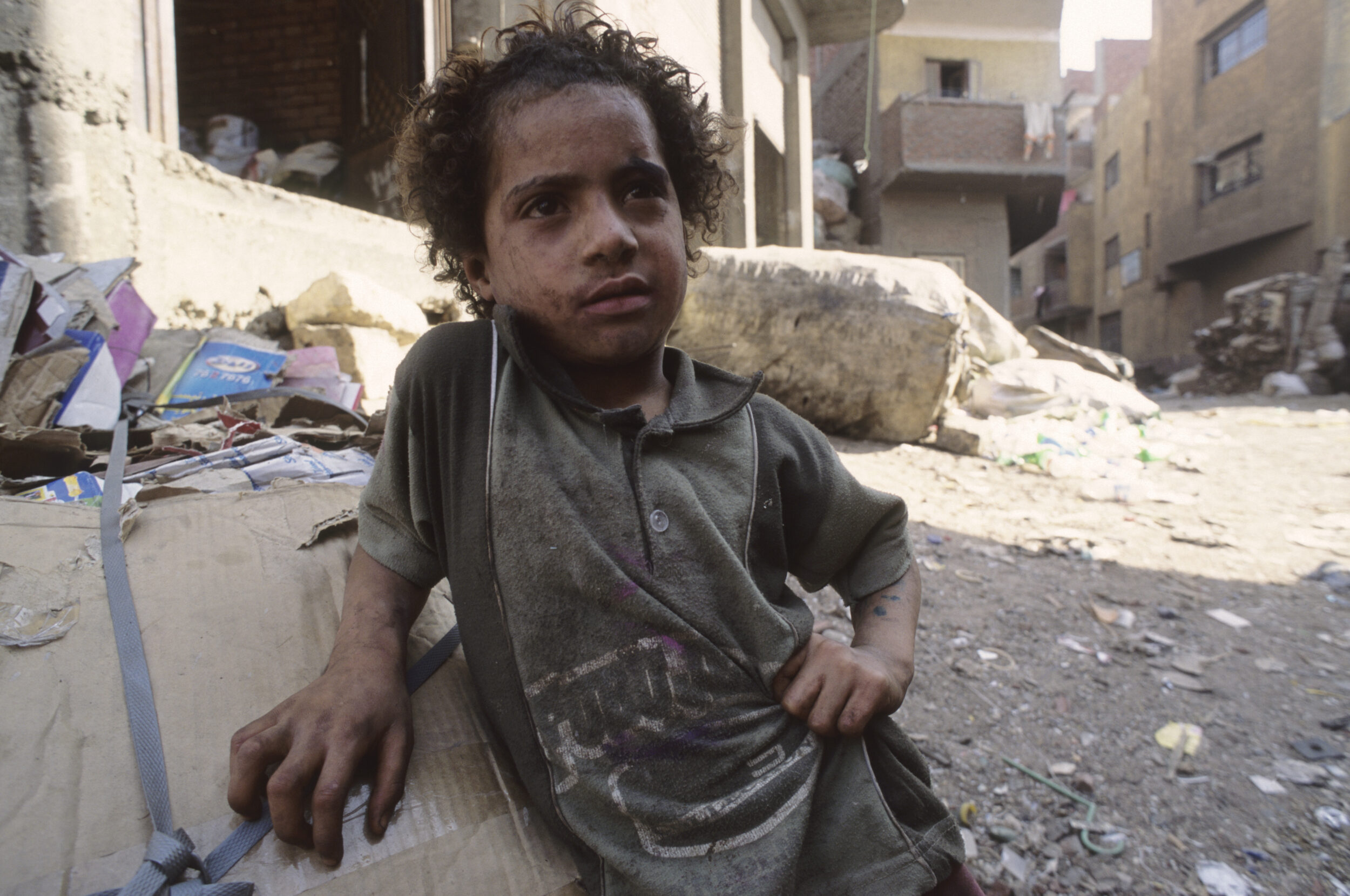Photo de portrait d'un jeune garçon chiffonnier au visage et aux mains très sale dans la rue, il est adossé à une balle de cartons triés, à l'ombre