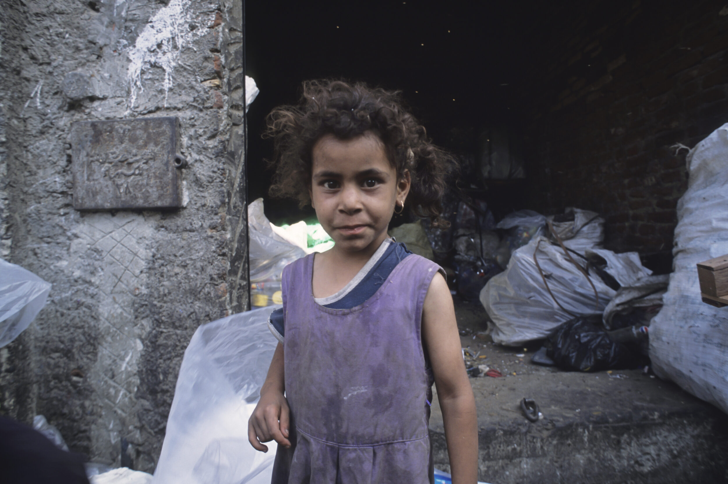 Portrait posé d'une enfant chiffonnière dans la rue devant la porte d'une maison entourée par des sacs de déchets, elle porte une robe violette et regarde dans l'objectif