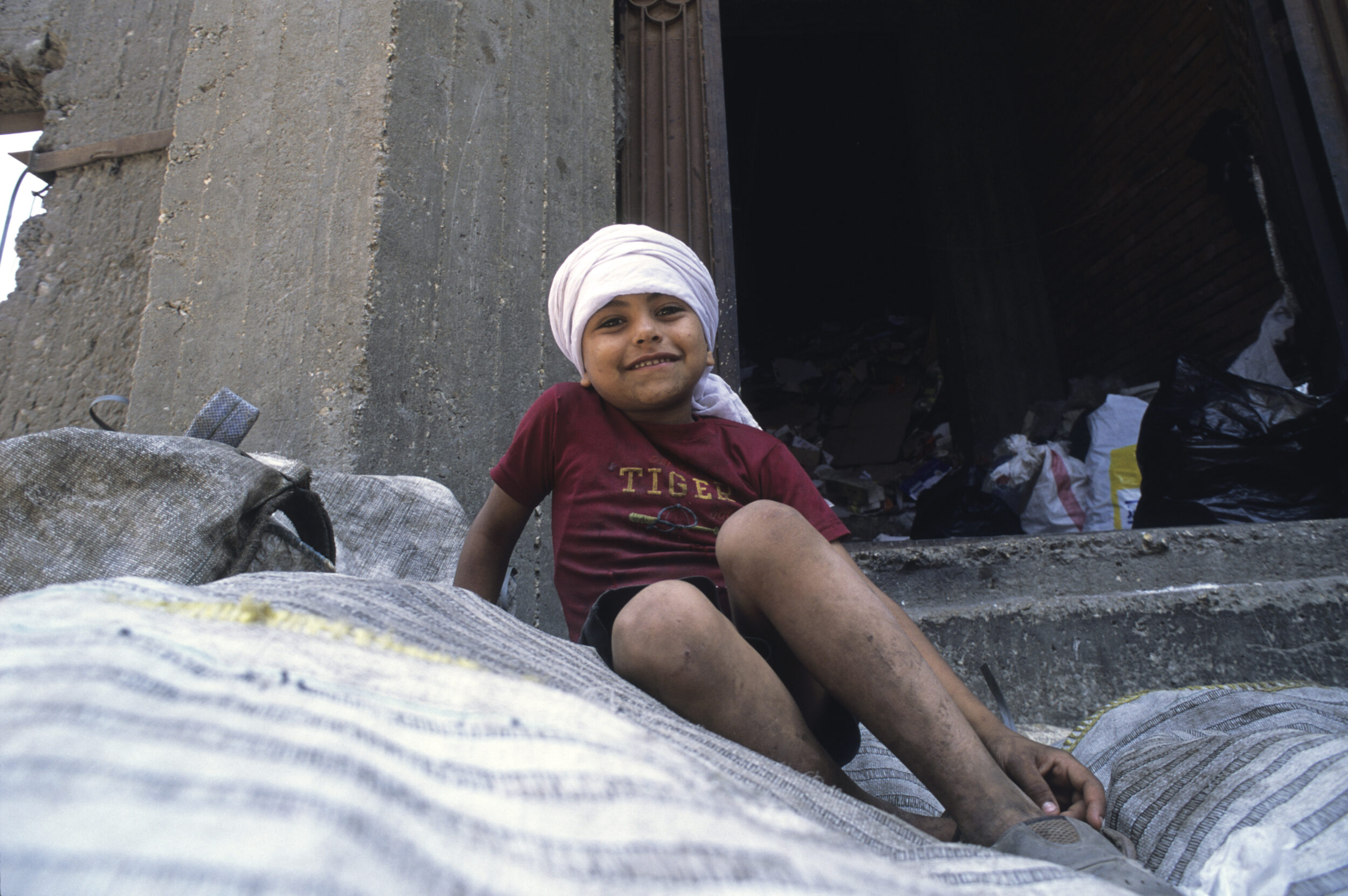 Portrait d'un jeune garçon Chiffonnier souriant portant un cheich, il est assis sur un tas de sacs remplis de déchets triés valorisables et regarde dans l'objectif