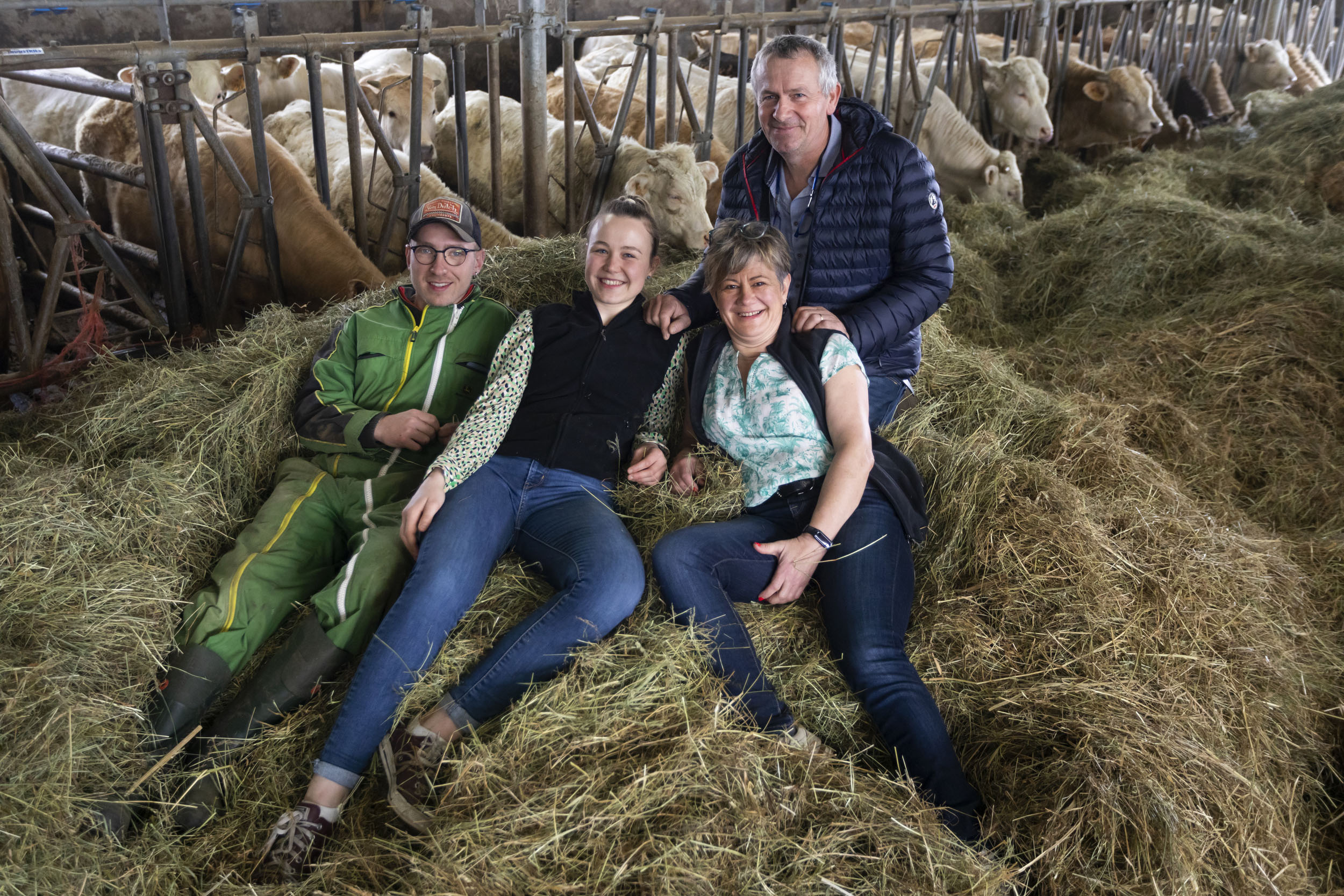 Portrait en plongée d'une famille d'éleveurs heureuse qui pose dans le foin au milieu de l'étable, les vaches sont à l'arrière plan