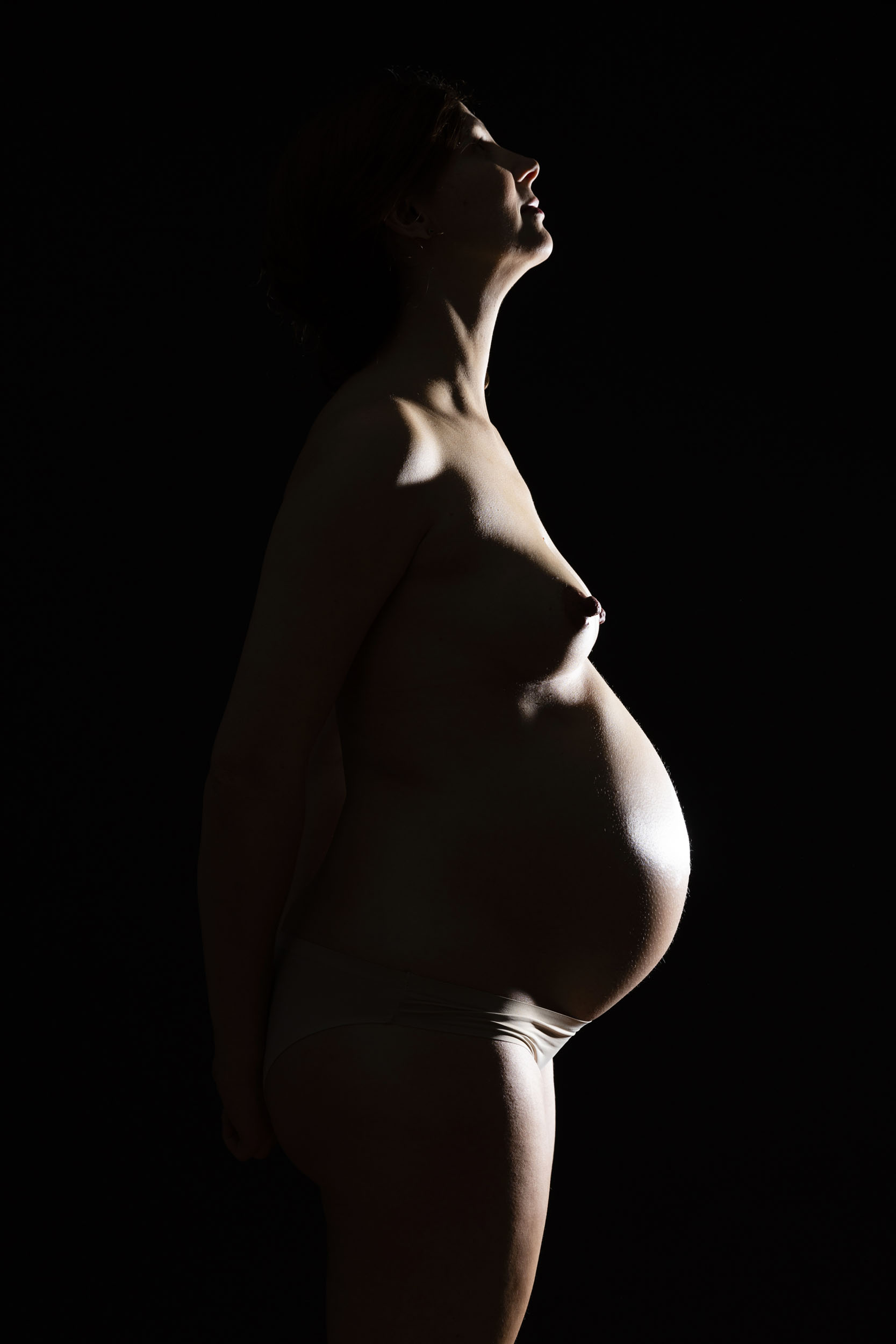 Portrait en pied d'une femme enceinte nue de profil en contre-jour sur fond noir, photographie réalisée en studio