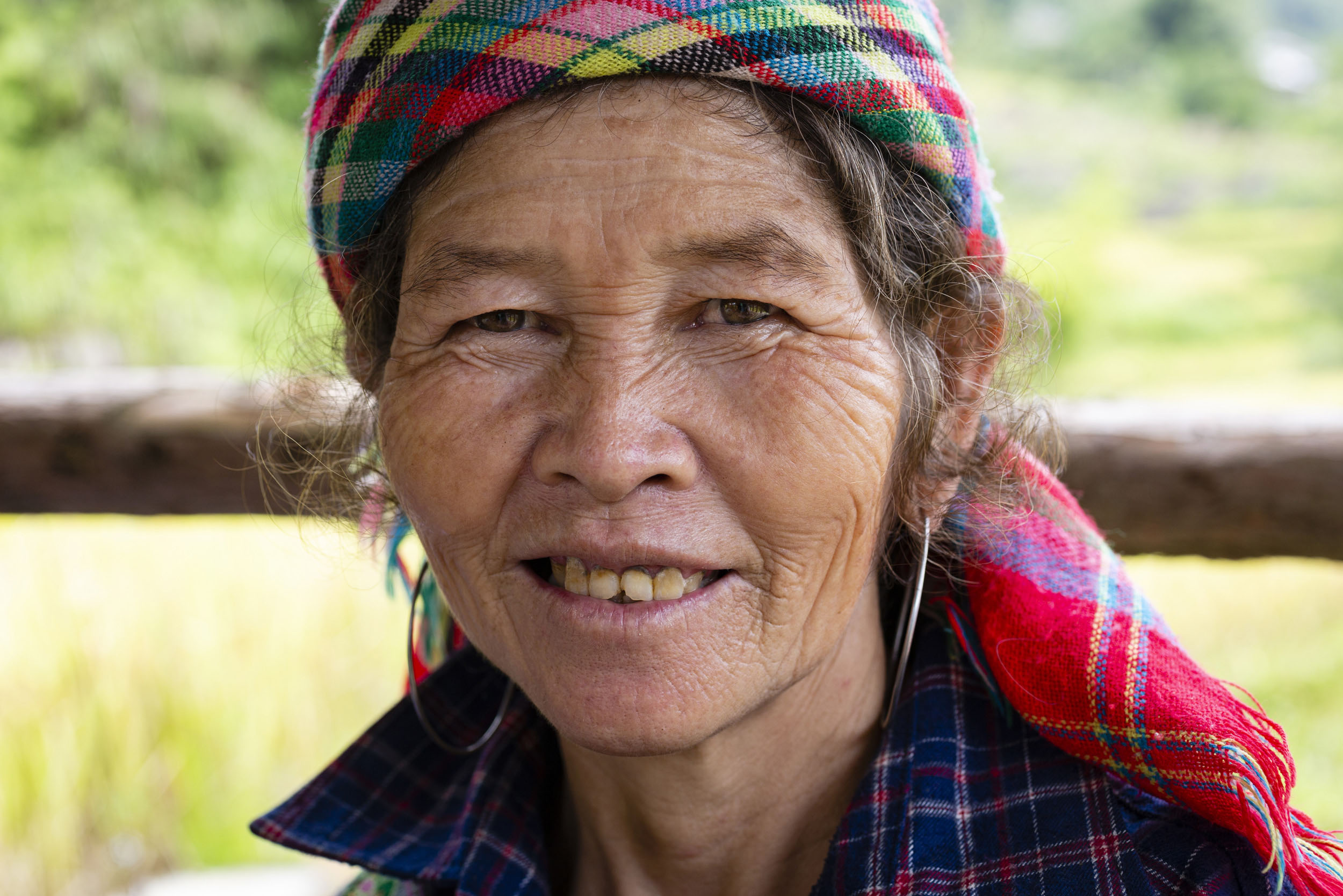 Portrait en gros plan d'une femme Vietnamienne ridée et souriante qui porte des anneaux en argent aux oreilles, un foulard sur la tête et regarde dans l'objectif. Derrière elle, on distingue un paysage de rizière flou