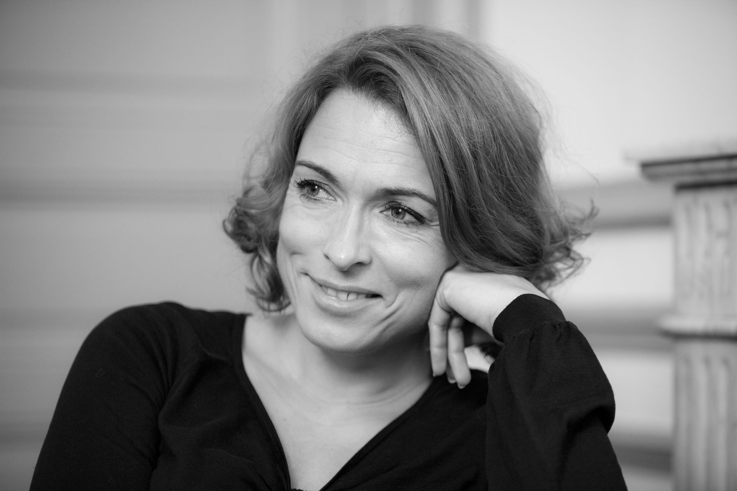 Portrait en noir et blanc d'une dirigeante souriante en réunion dans un salon haussmannien, elle regarde son interlocuteur. Une cheminée en marbre est floue à l'arrière plan