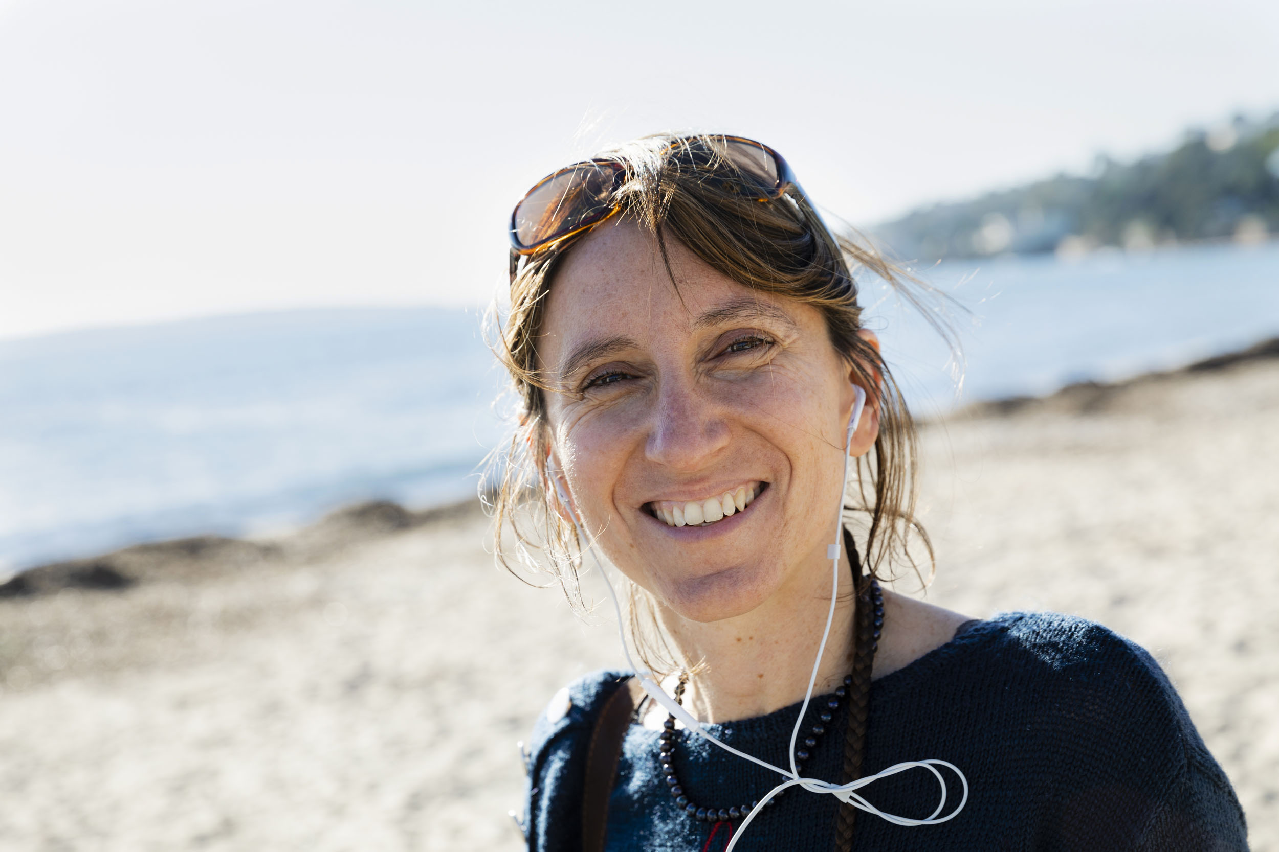 Portrait d'une femme souriante en pull bleu sur une plage en hiver avec ses écouteurs et ses lunettes de soleil sur la tête, elle regarde dans l'objectif
