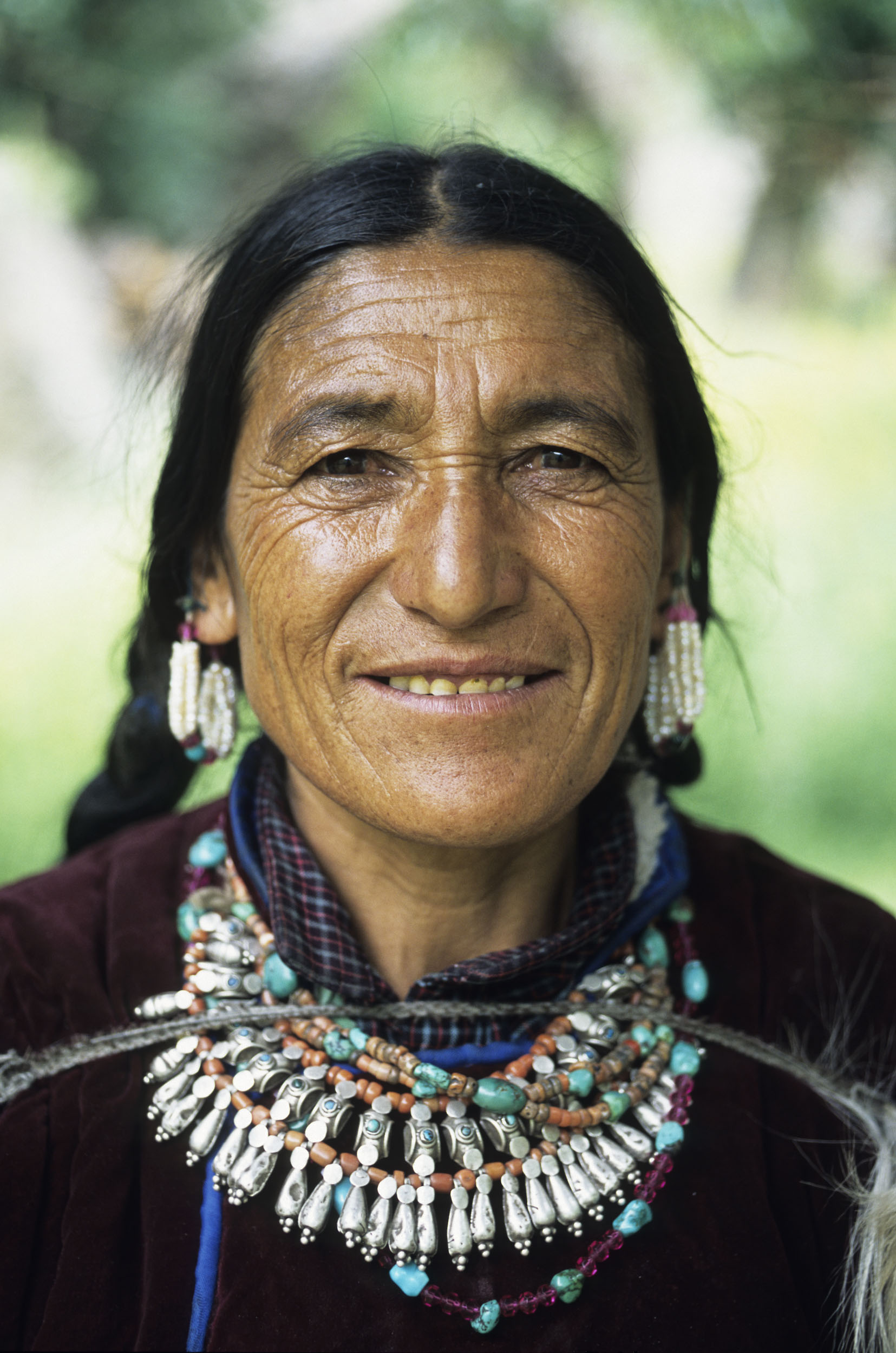 Portrait posé en gros plan d'une vieille femme Ladakhi ridée et souriante aux cheveux brun et longs, elle est en costume traditionnel et porte de nombreux bijoux