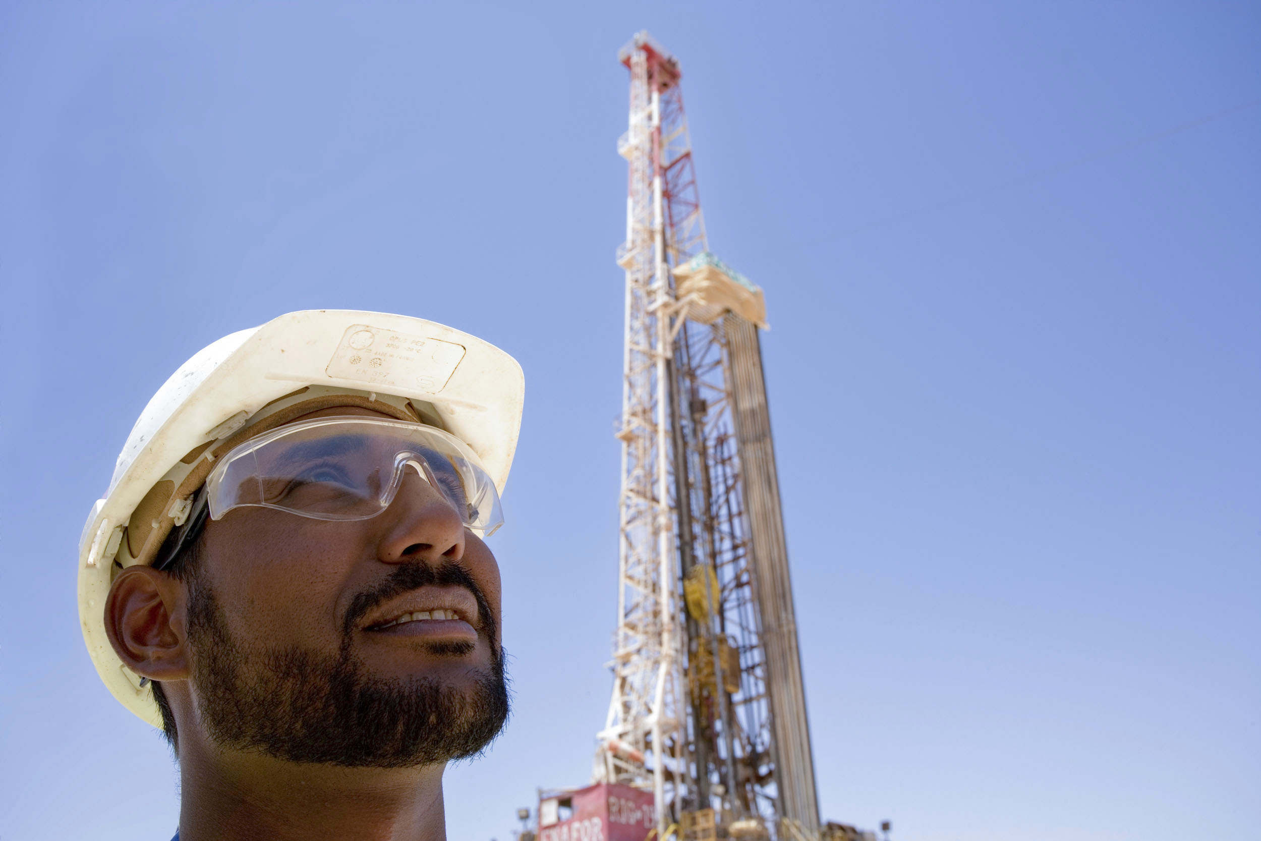 Portrait d'un foreur en contre-plongée devant un rig de forage qui porte un casque blanc et des lunettes de protection, il regarde loin devant lui vers le haut, le ciel est bleu