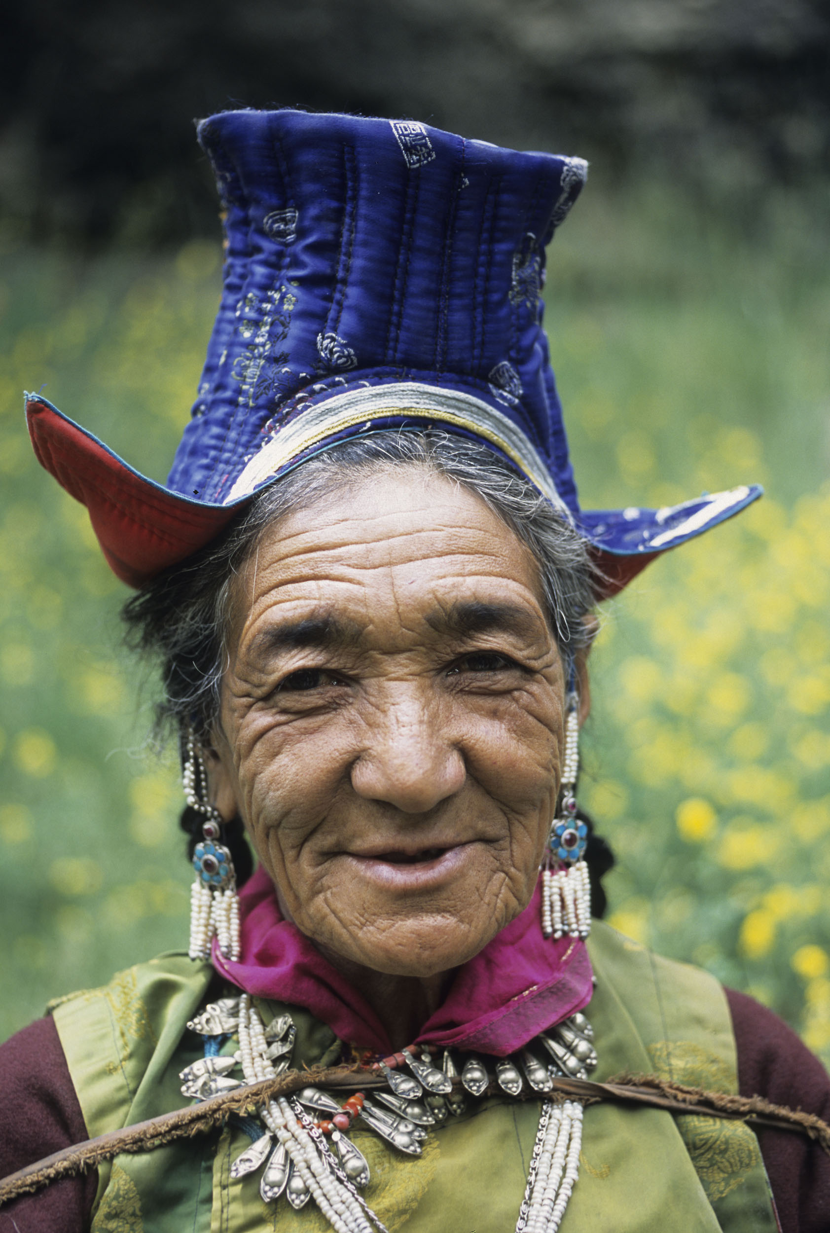 Portrait posé en gros plan d'une vieille femme Ladakhi ridée et souriante aux cheveux brun et longs, elle est en costume traditionnel et porte de nombreux bijoux