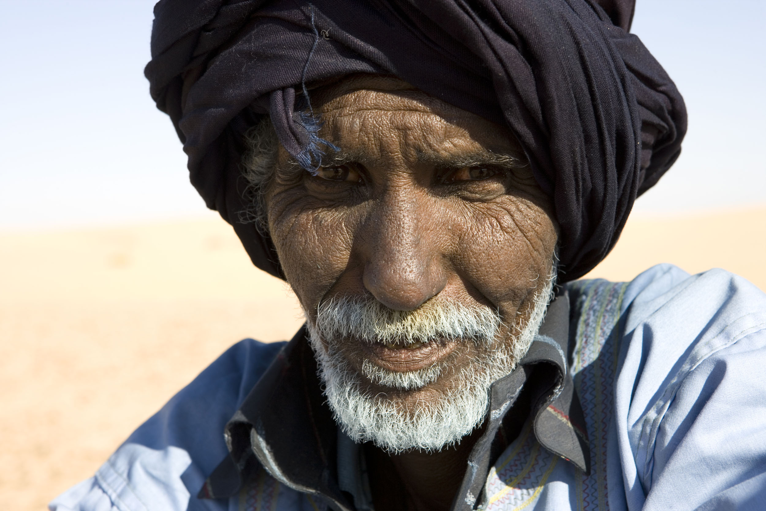 Portrait de face en gros plan du visage d'un homme mauritanien dans le désert qui regarde dans l'objectif, il porte un cheich bleu foncé noué sur la tête et une djellaba bleu ciel