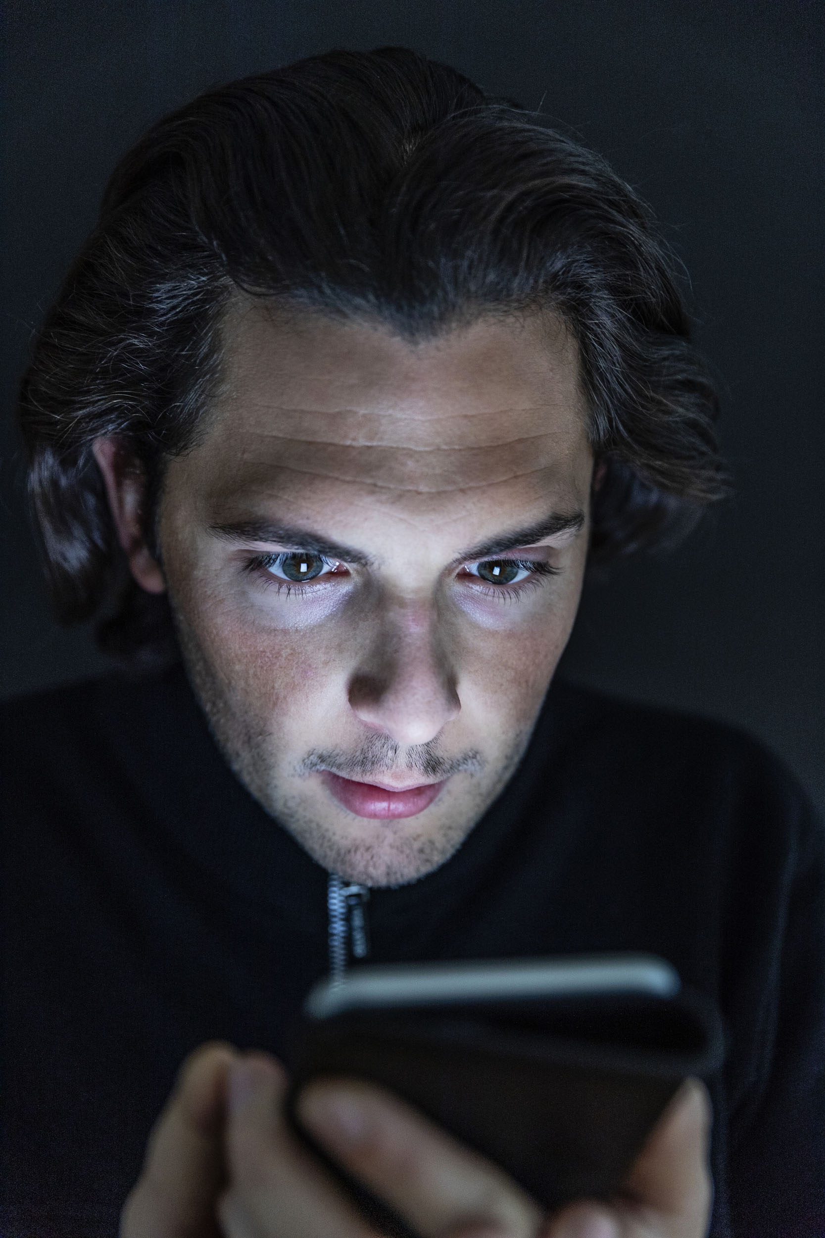 Portrait d'un homme en gros plan au visage éclairé par l'écran de son téléphone portable par lequel il est totalement absorbé, ses yeux sont rivés sur l'écran