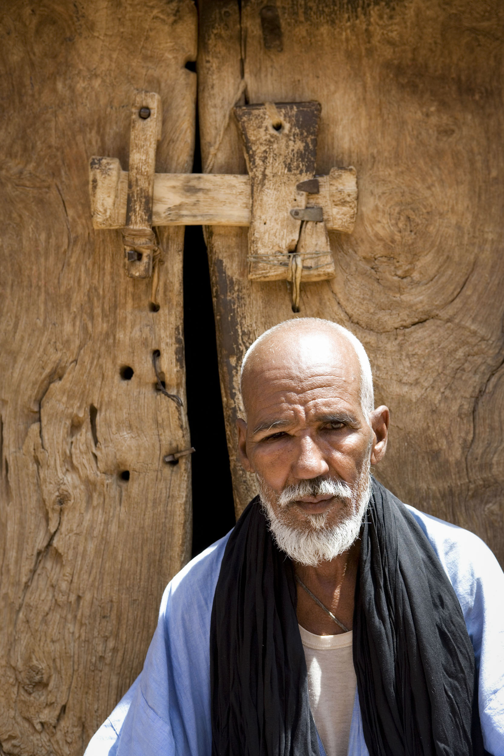 Portrait posée d'un homme mauritanien portant barbe et moustache devant la porte en bois de sa maison, il est vêtu d'une djellaba bleue et d'un cheich noir autour du cou