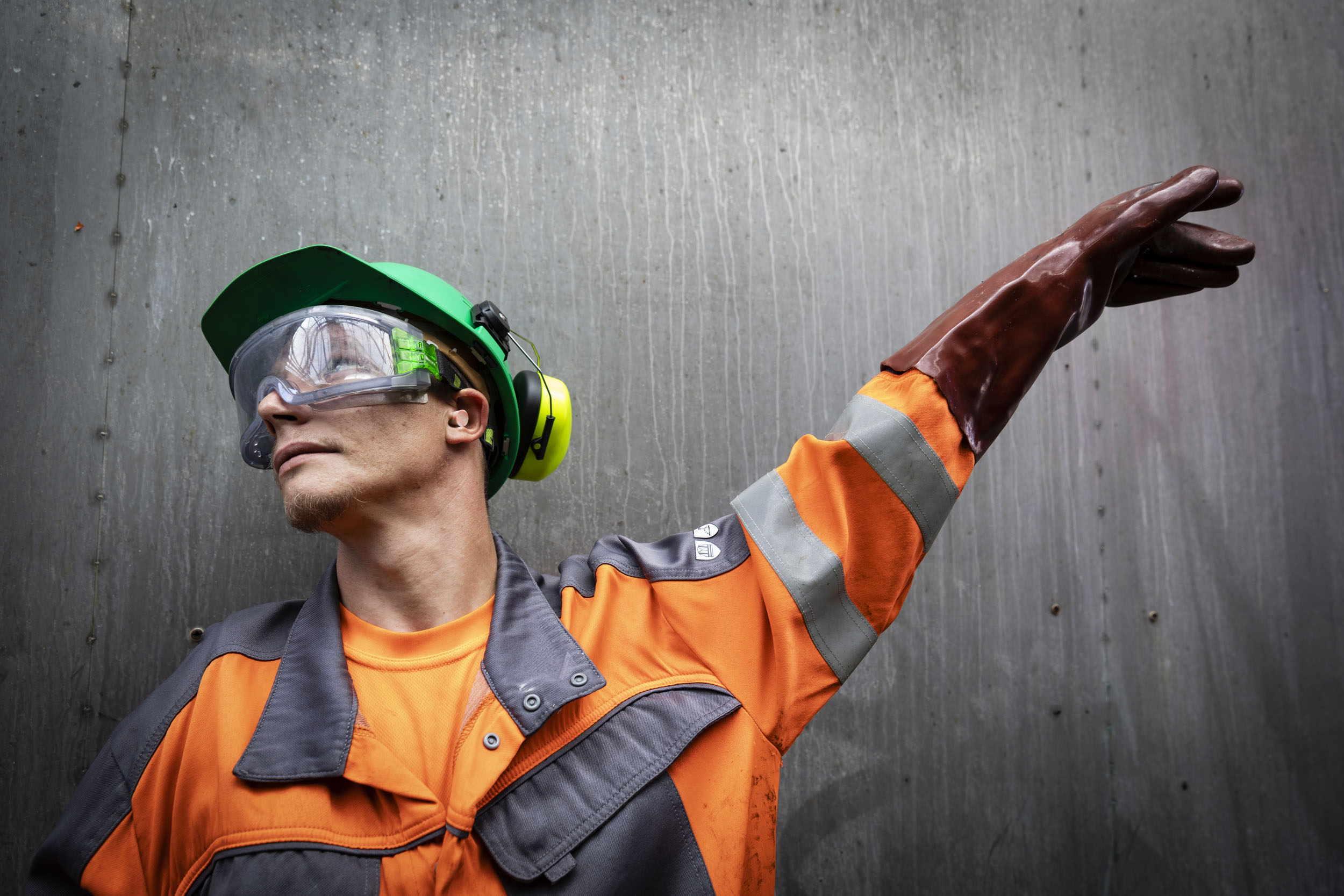 Portrait en contre-plongée d'un homme debout contre un mur dans une station de lavage industrielle de camion citerne. Il porte une combinaison de travail orange et grise, un casque vert, un casque anti bruits et des gants de protection épais en caoutchouc