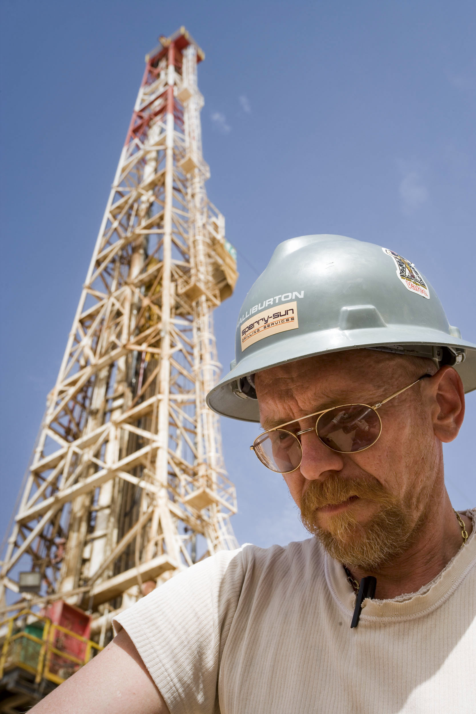 Portrait en contre plongée d'un foreur au travail au pied d'un rig de forage sur fond de ciel bleu. Il porte un casque vert, des lunettes de soleil et un T-Shirt blanc usé
